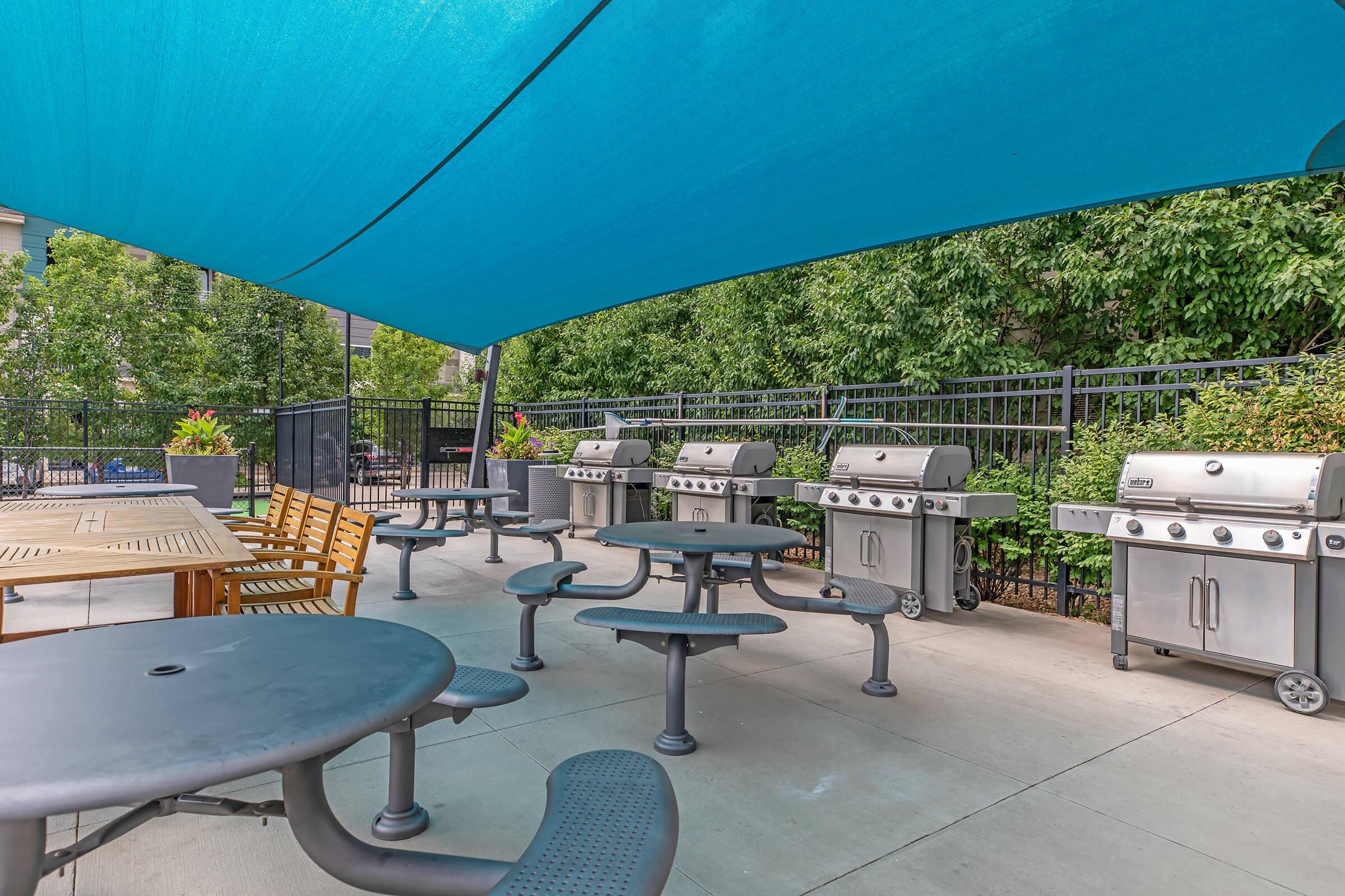 Outdoor grilling area featuring multiple metal grills under a blue shade canopy. There are wooden tables and benches, surrounded by greenery and a black fence, creating a comfortable space for outdoor cooking and dining. The setting is clean and inviting for gatherings.