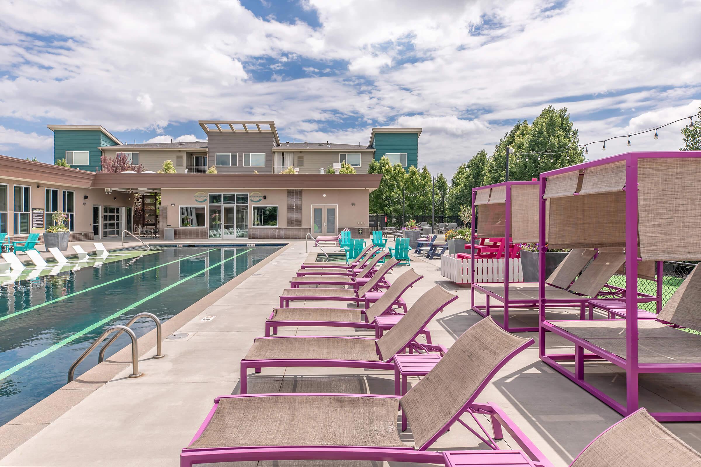 A resort-style swimming pool surrounded by vibrant pink lounge chairs and cabanas. The pool area features modern outdoor furniture and a backdrop of a stylish building with large windows. Lush greenery and a partly cloudy sky complete the relaxing atmosphere.