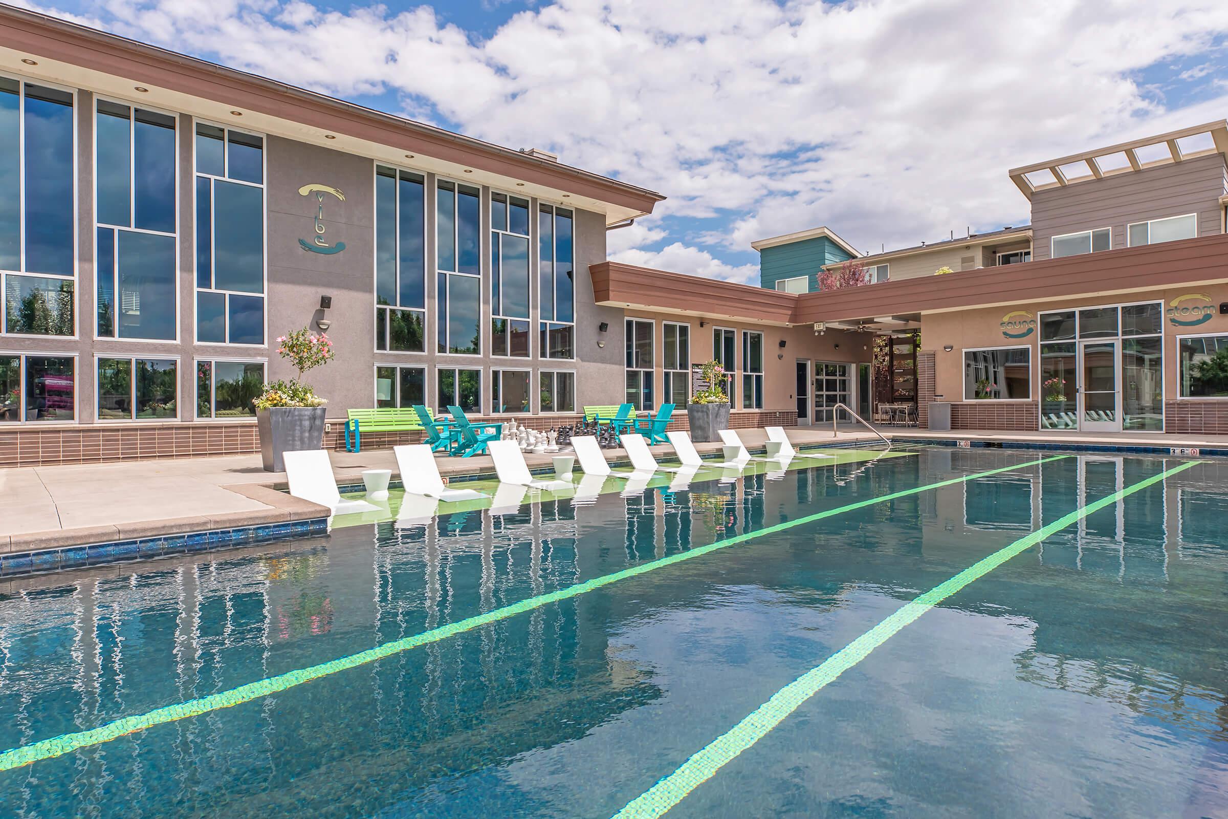 A modern swimming pool area featuring clear blue water, surrounded by lounge chairs and an elegant building with large windows. The sky is partly cloudy, and there are potted plants adding greenery to the setting, creating a relaxing and inviting atmosphere.