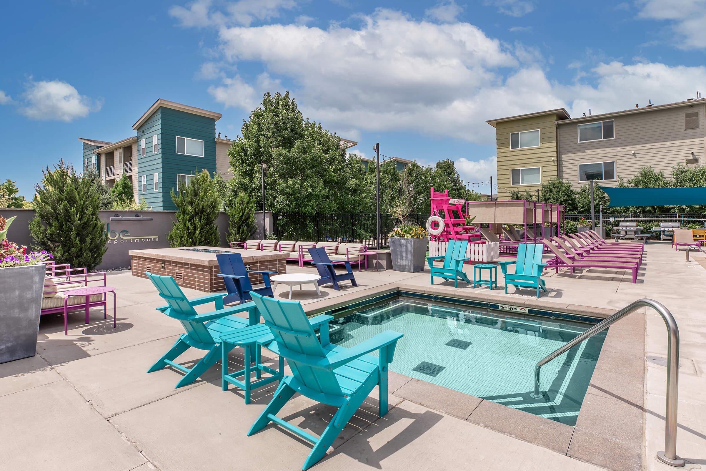 A vibrant pool area featuring colorful lounge chairs in shades of pink and blue. In the foreground, a hot tub is surrounded by chairs, while the background showcases modern apartment buildings and lush greenery under a clear blue sky.
