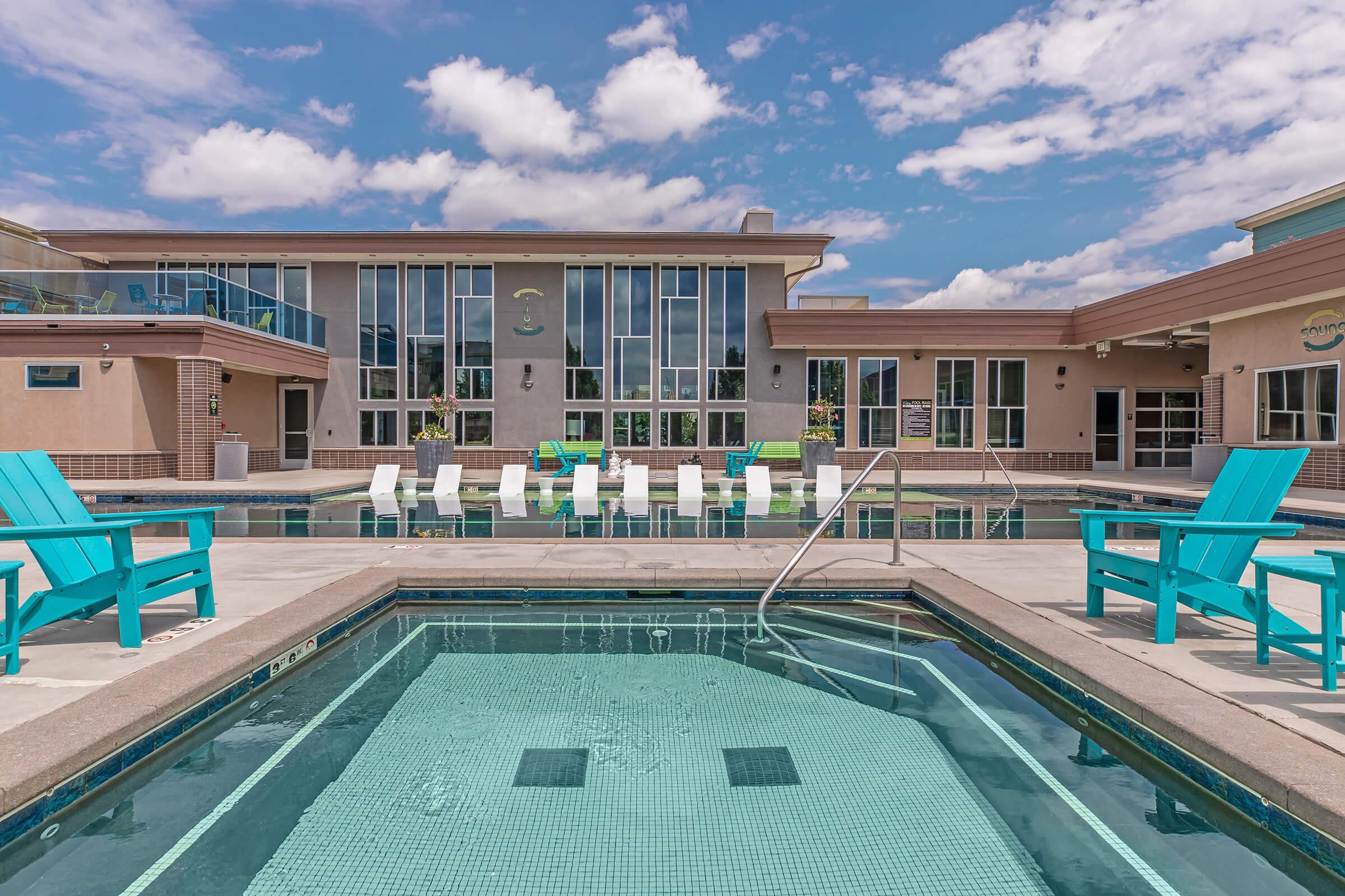 Outdoor swimming pool area with clear blue water, surrounded by light-colored buildings and lounge chairs. A separate hot tub is visible in the foreground. The sky is partly cloudy, creating a bright and inviting atmosphere for relaxation.