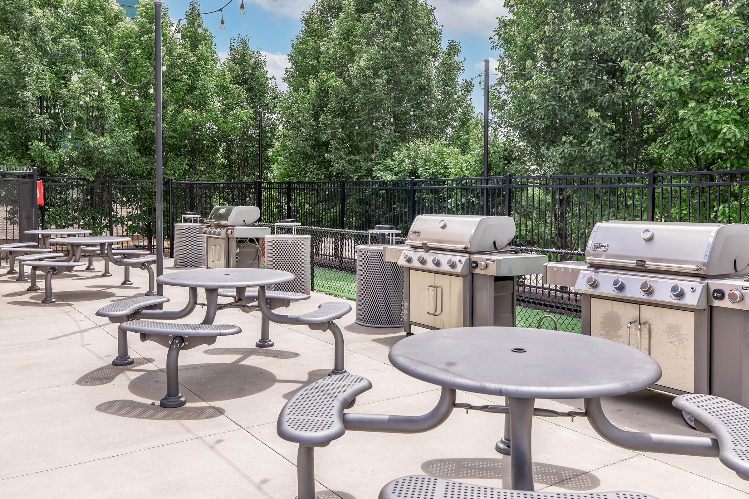 A communal outdoor grilling area featuring multiple barbecue grills and several picnic-style tables, surrounded by green trees and a fence, with clear skies in the background.