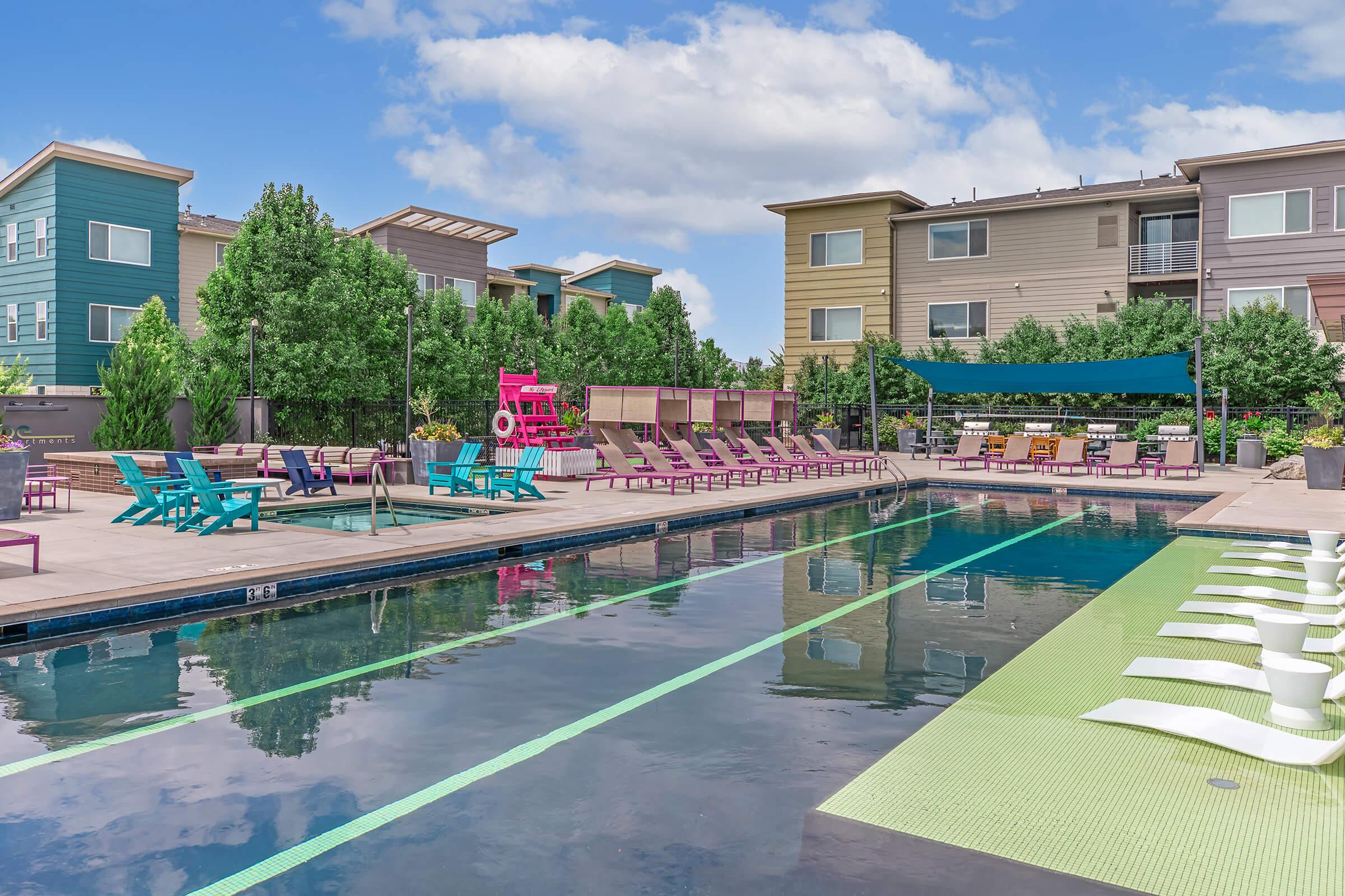 A modern swimming pool area surrounded by colorful lounge chairs and greenery. The pool features lanes for swimming, and there are shaded seating areas with vibrant canopies. The backdrop includes contemporary-style apartment buildings under a partly cloudy sky.