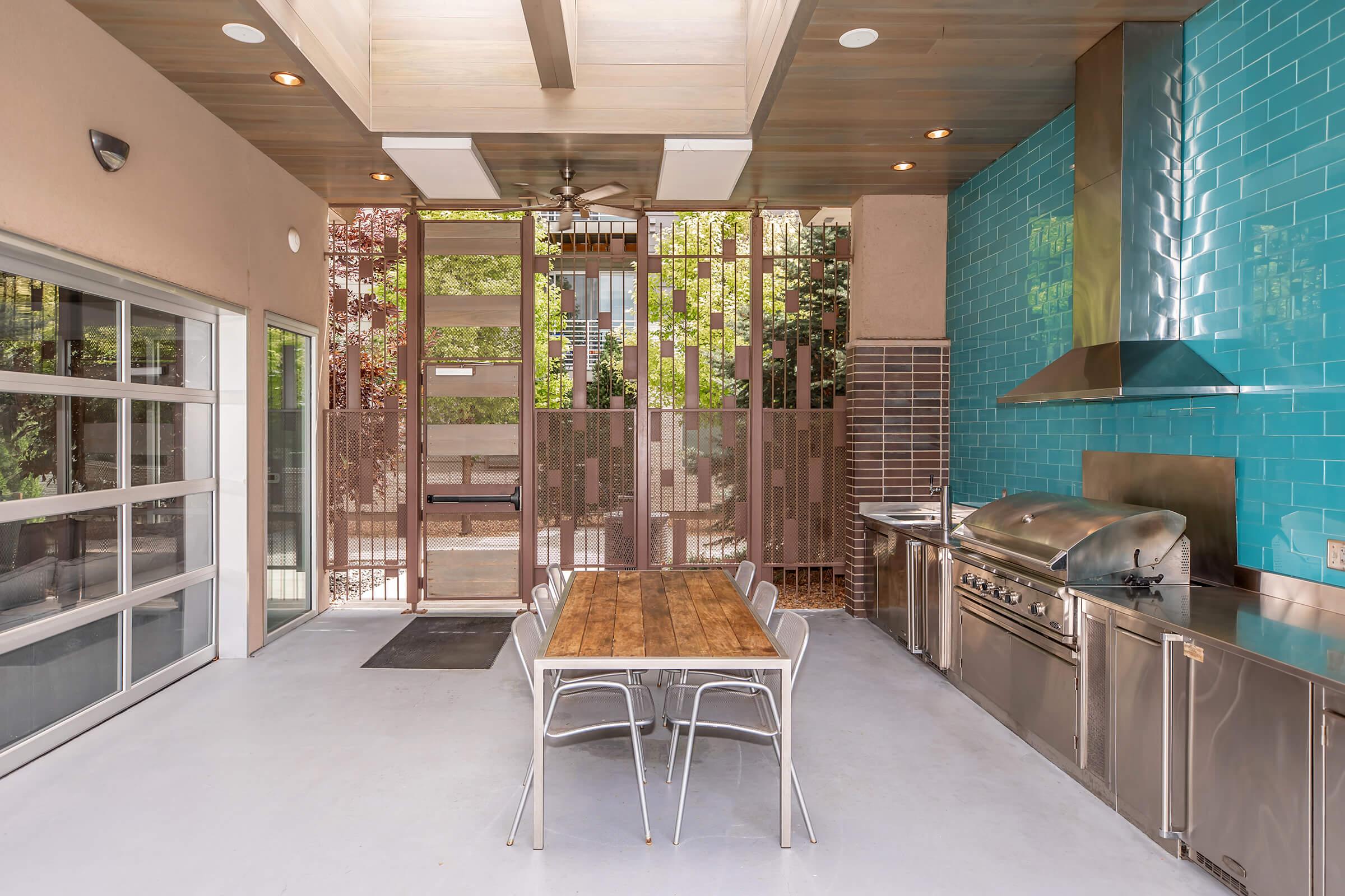 An outdoor kitchen area features a stainless steel grill, modern appliances, and a wooden dining table with metal chairs. The space is brightened by large windows and is complemented by blue tiled walls and decorative screens, creating a contemporary and inviting atmosphere.