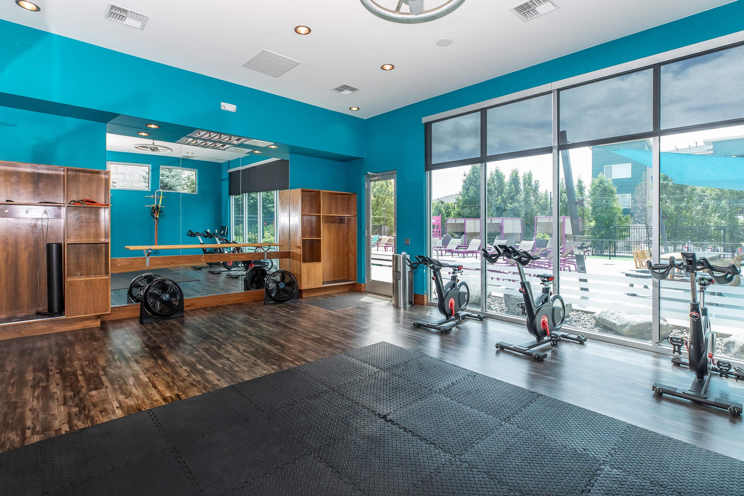 Modern gym interior with vibrant blue walls, wooden storage cabinets, and large windows providing natural light. The space features exercise equipment, including stationary bikes and weights, arranged on a dark rubber floor, creating an inviting workout environment.
