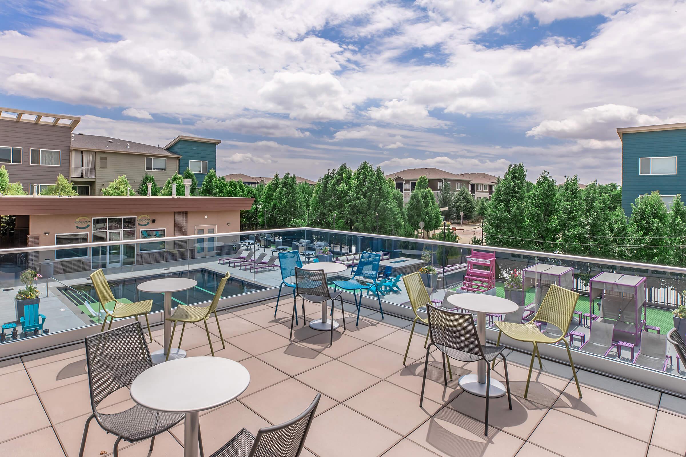 Rooftop terrace with colorful lounge chairs and tables overlooking a pool and landscaped area. In the background, modern apartment buildings and trees are visible under a partly cloudy sky.