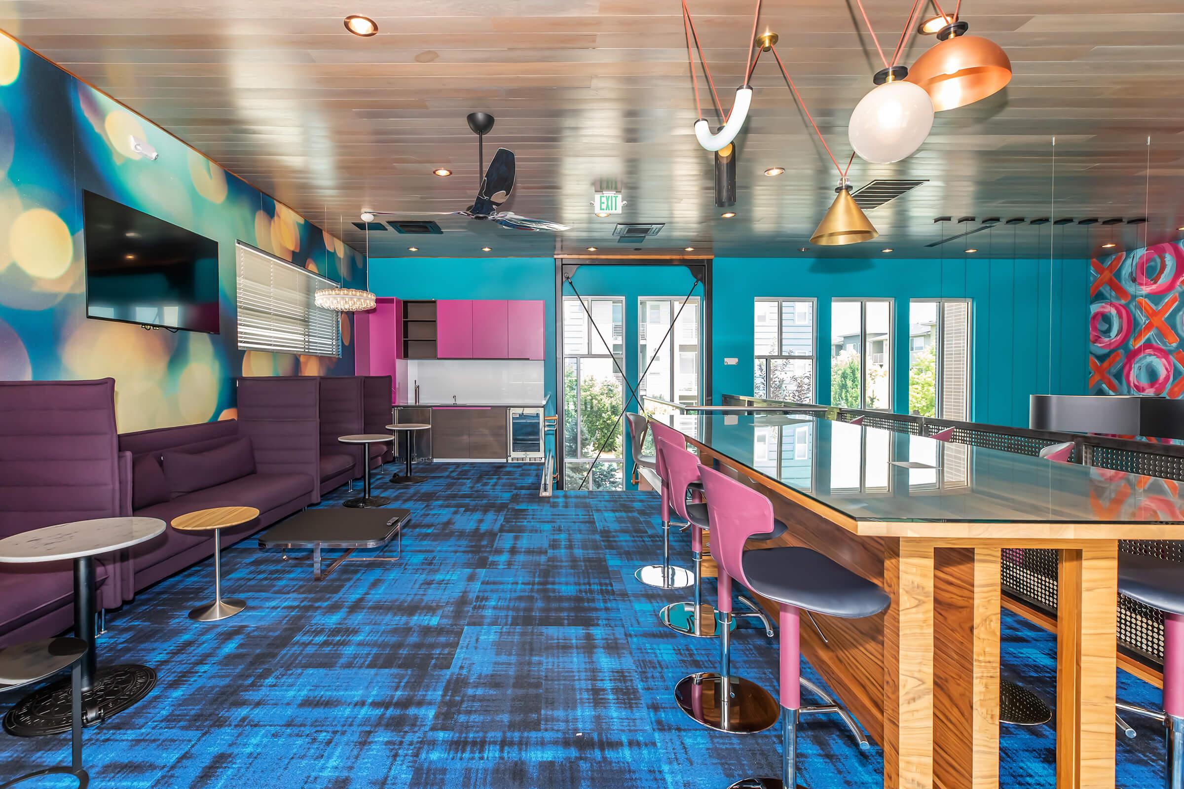 A modern lounge area featuring vibrant blue carpet, colorful walls with abstract patterns, and stylish lighting. The room includes a sleek bar with purple and wooden stools, cozy seating areas with purple couches, and a flat-screen TV. Large windows provide natural light and views of the outdoors.