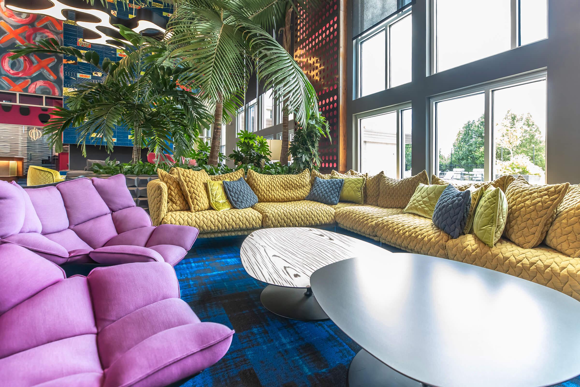 A modern lounge area featuring a large, textured yellow sofa and several purple armchairs. The space is filled with natural light from large windows, surrounded by greenery. A stylish round coffee table sits in the center on a vibrant blue carpet, creating a cozy and inviting atmosphere.