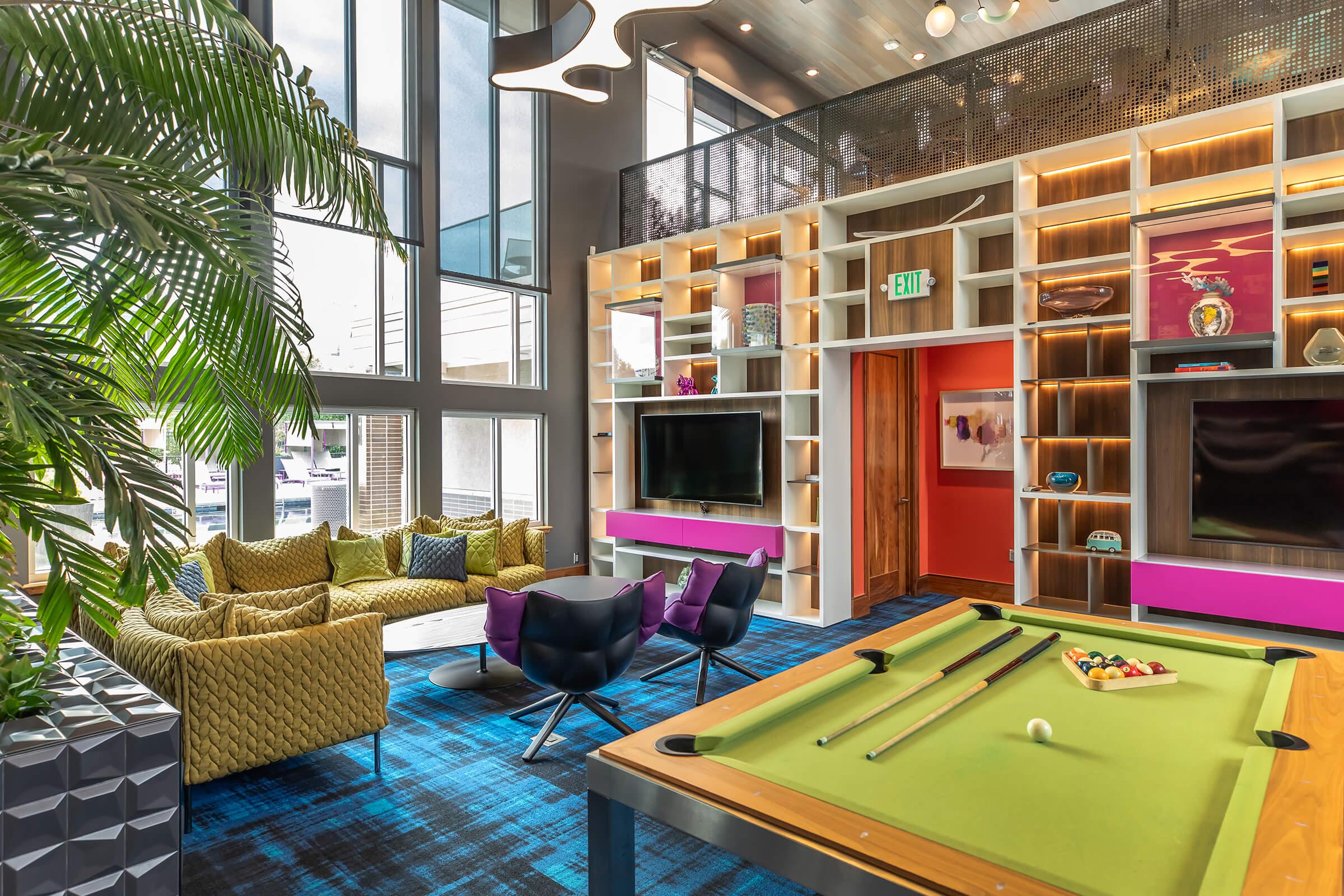 A modern lounge area featuring a billiards table, plush seating, and large windows. The decor includes a vibrant color palette with purple and yellow accents, and there's a stylish bookshelf displaying decorative items. A sense of openness and contemporary design is evident throughout the space.