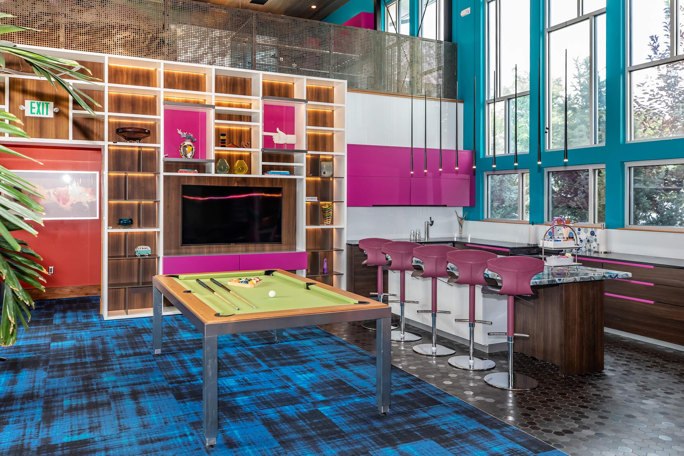 A bright and modern recreational room featuring a green pool table, a large wall-mounted TV, and a stylish bar area with purple stools. The space is adorned with colorful shelving and large windows, letting in plenty of natural light, against a blue patterned carpet.