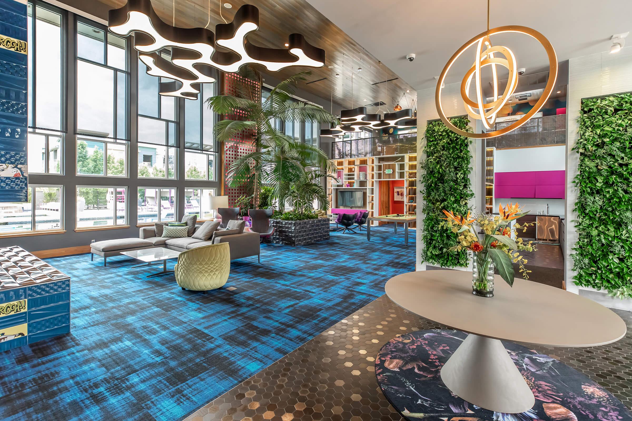 A modern lobby area with large windows, featuring stylish furniture in vibrant colors and patterns. The space includes decorative plants, unique light fixtures, and a circular table adorned with flowers, creating a welcoming and contemporary atmosphere.