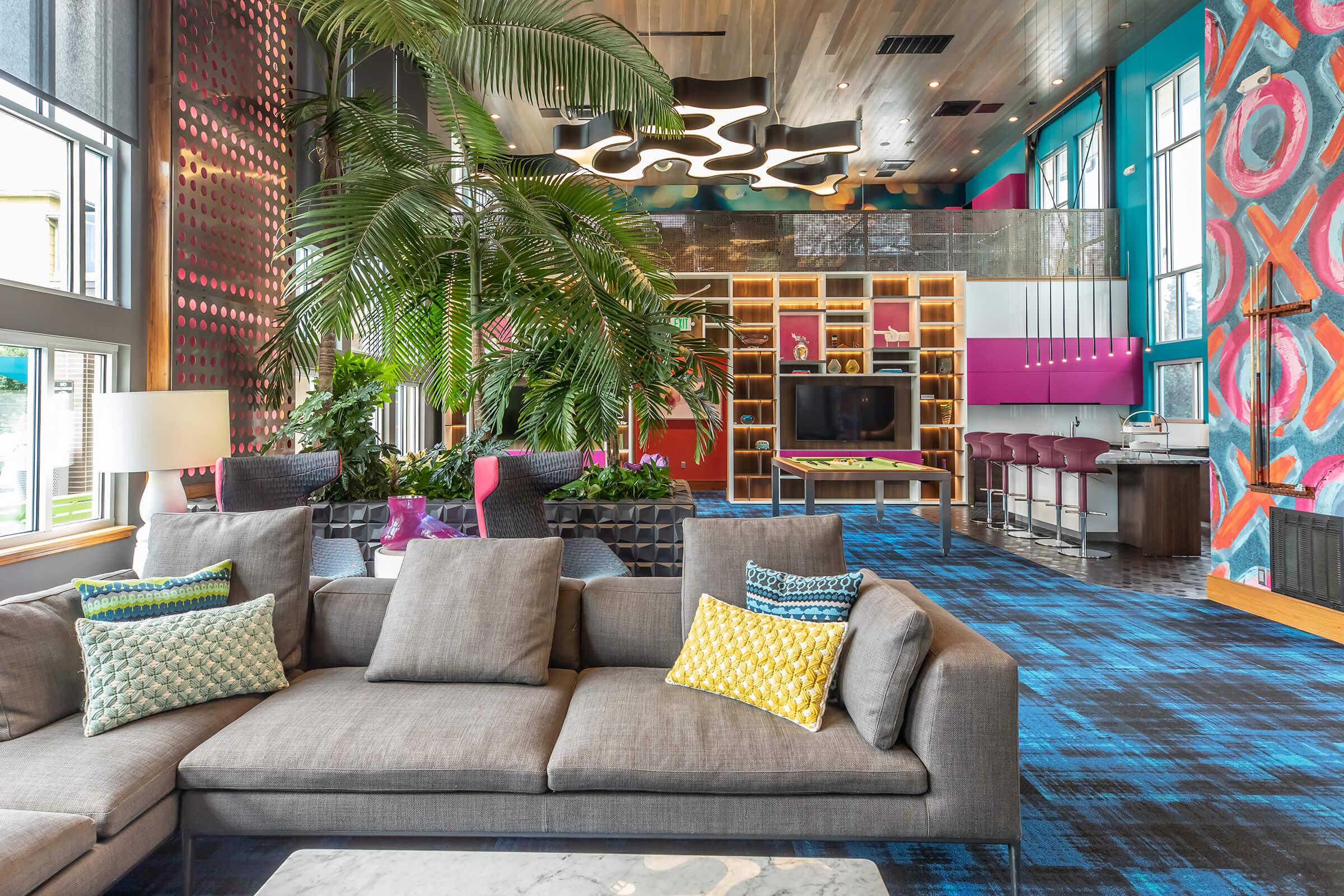 A modern lounge area featuring a stylish gray sofa adorned with colorful throw pillows. The space includes vibrant walls with abstract patterns, lush indoor plants, and a sleek kitchen area with bar stools and a TV. Large windows allow natural light to fill the room, creating a welcoming atmosphere.