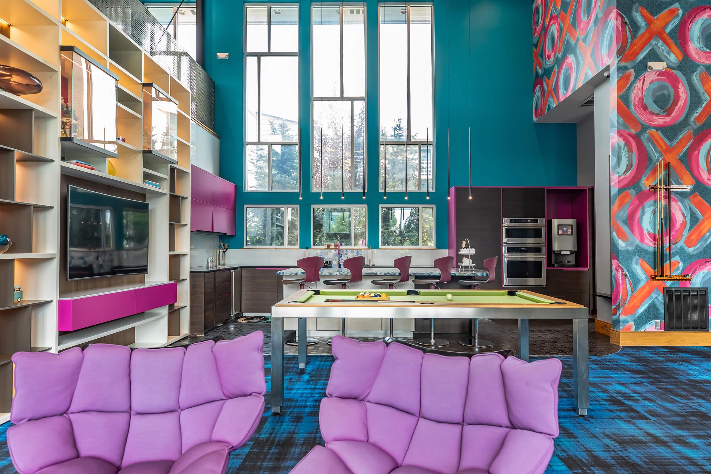 A modern and colorful interior of a spacious kitchen and living area. Features vibrant turquoise walls, large windows, and a stylish open layout. The space includes purple lounge chairs, a dining table, a contemporary kitchen with sleek appliances, and a bold patterned accent wall, creating a lively atmosphere.