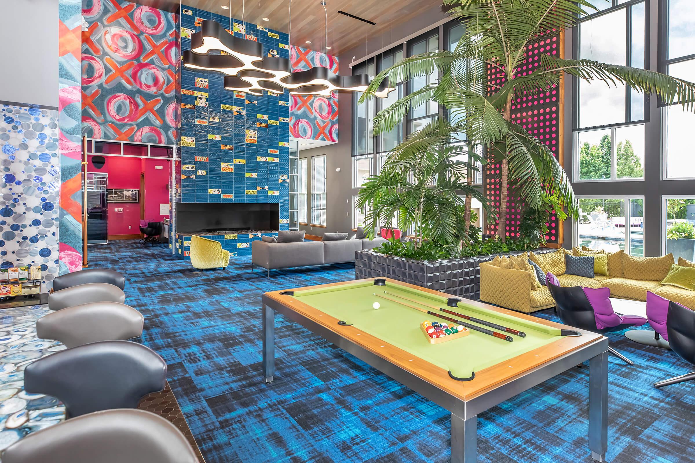 Modern interior of a vibrant lounge area featuring a pool table, colorful carpet, and large windows. The space includes stylish seating with bright cushions, a wall of patterned tiles, indoor plants, and a cozy atmosphere, ideal for socializing and relaxation.