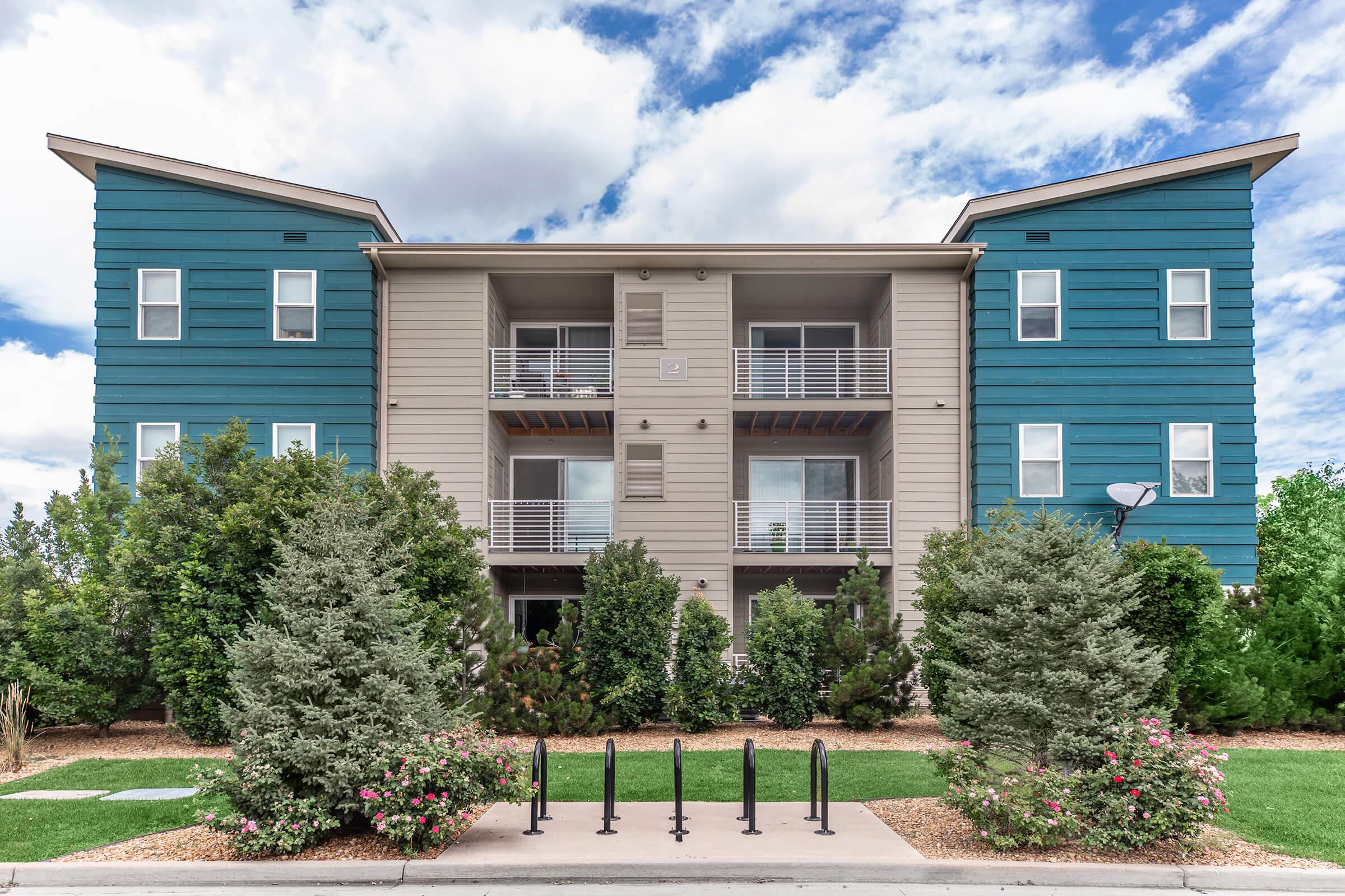 A modern three-story apartment building featuring a combination of blue and beige siding, surrounded by green landscaping. The front has multiple balconies and large windows, with flowering shrubs and trees in the foreground, under a partly cloudy sky.