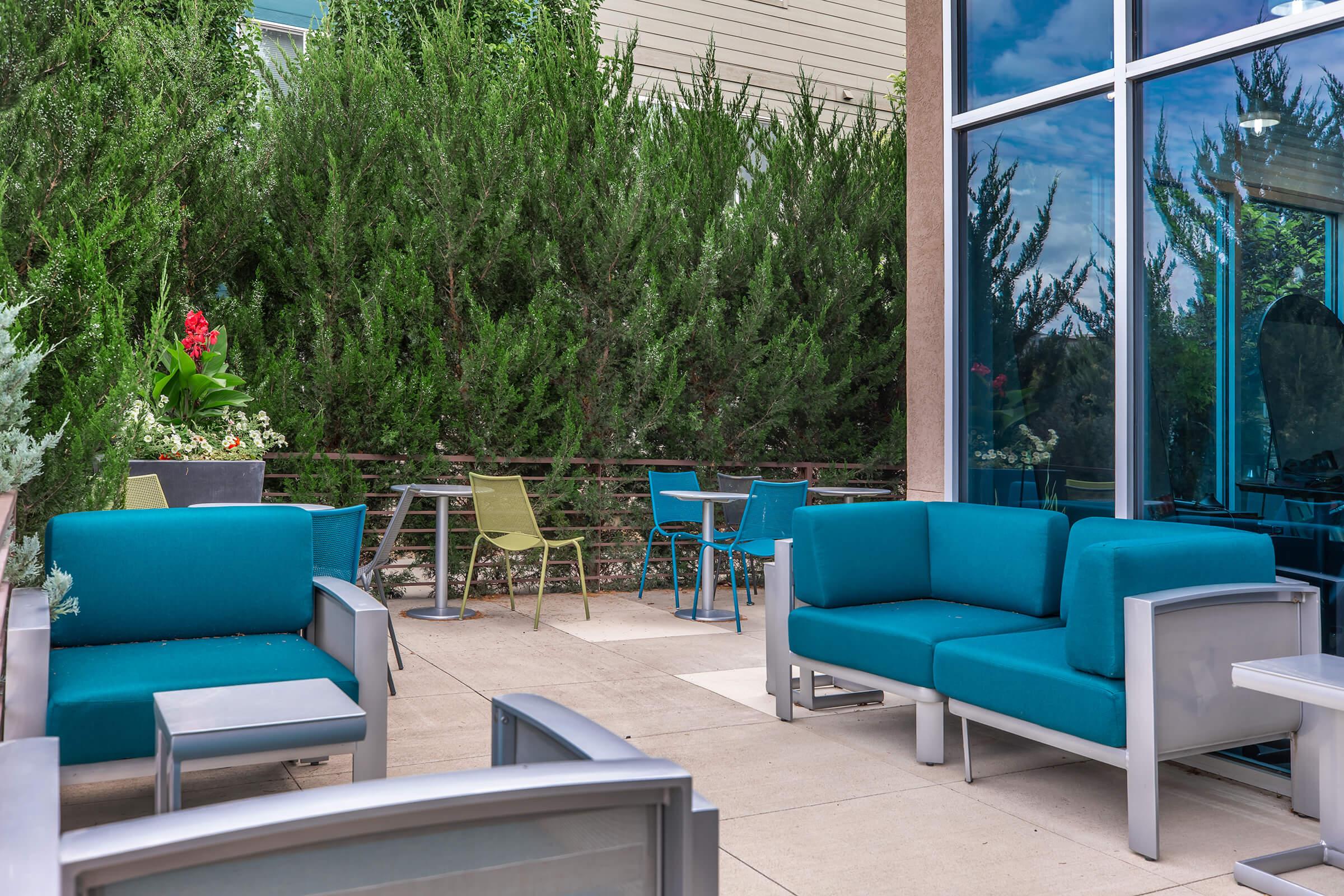 A cozy outdoor seating area featuring turquoise couches and chairs arranged around small tables. Lush greenery and a bright blue glass wall create a relaxing atmosphere, perfect for socializing or enjoying a peaceful moment outdoors.