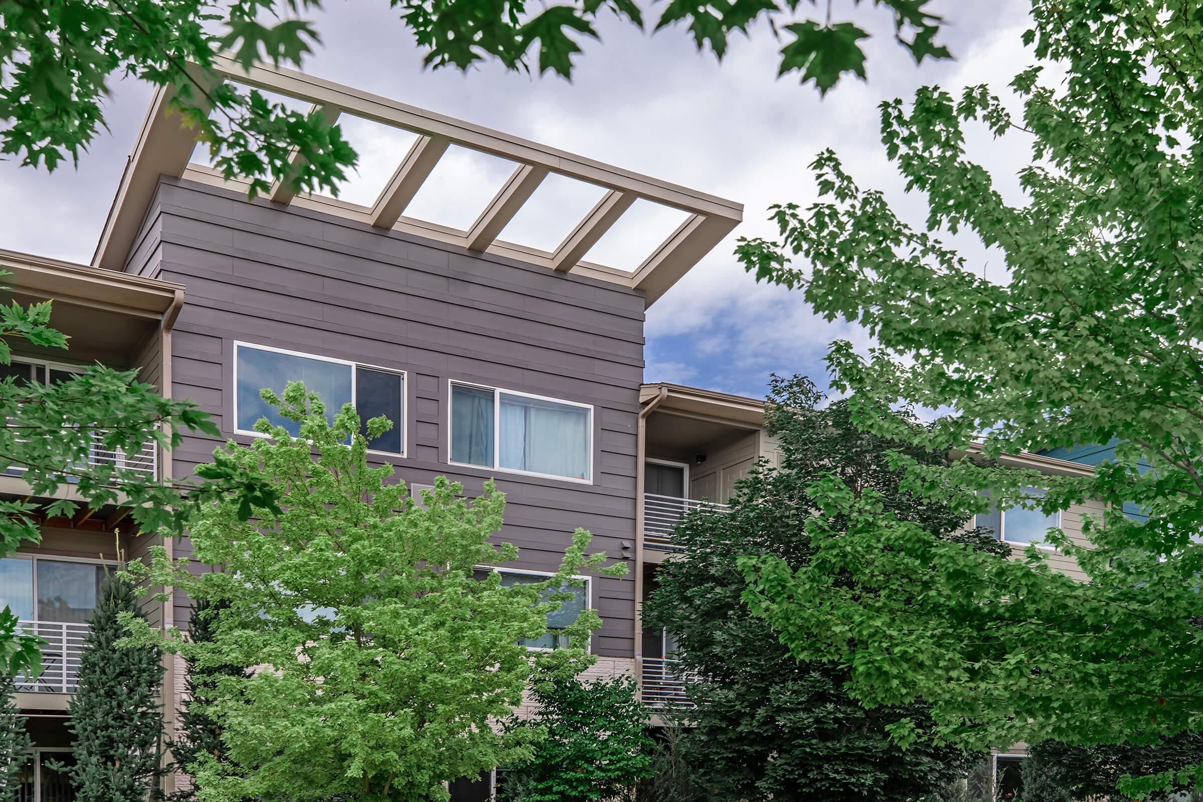 A modern multi-story building partially obscured by lush green trees. The structure features large windows and a distinctive overhanging roof design, against a backdrop of a cloudy sky. The blend of nature and architecture creates a serene urban scene.