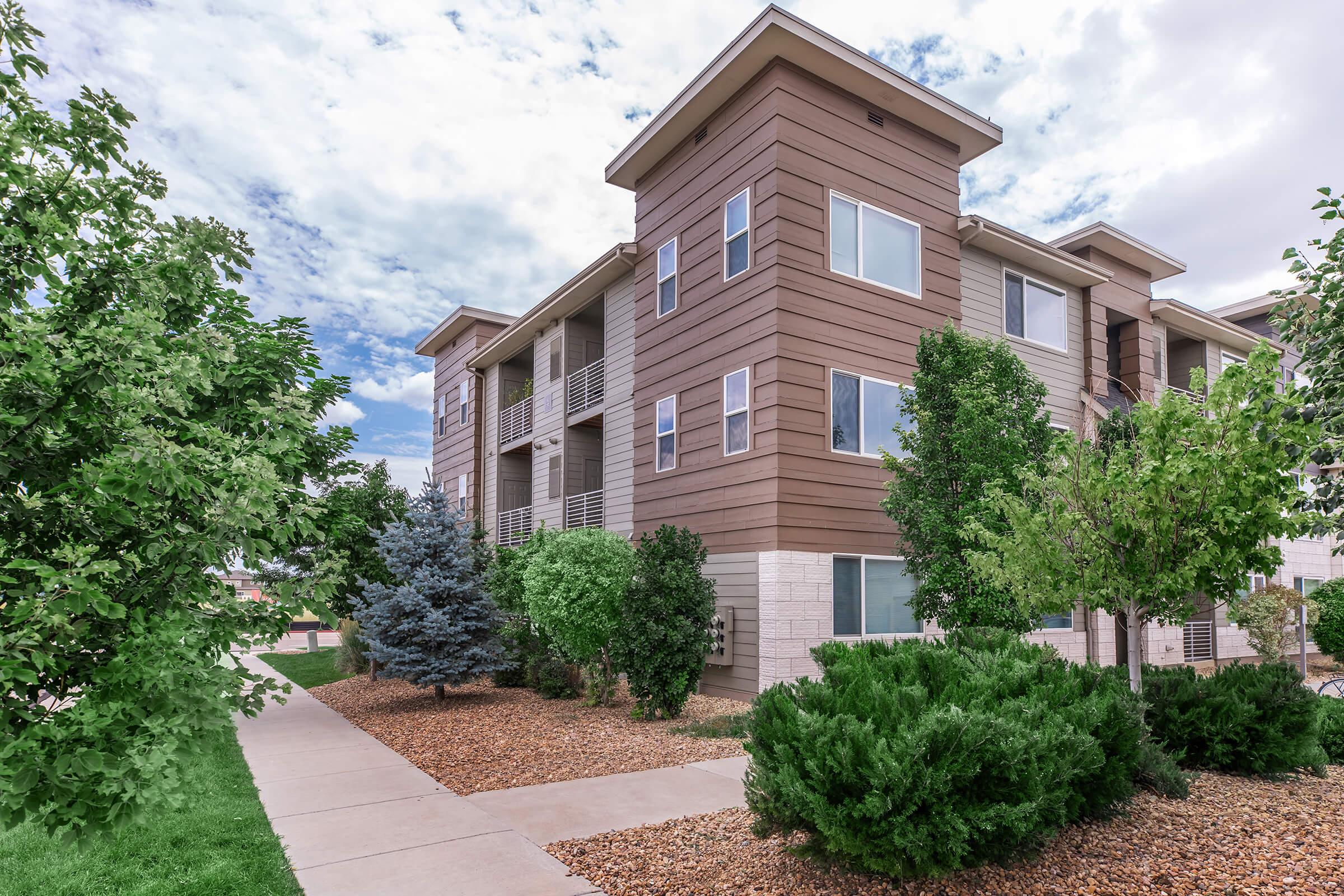 Modern multi-story apartment building with a mix of brown and beige siding. Surrounding greenery includes small trees and shrubs, with a pathway leading to the entrance. The sky is partly cloudy, adding to the inviting atmosphere of the residential area.