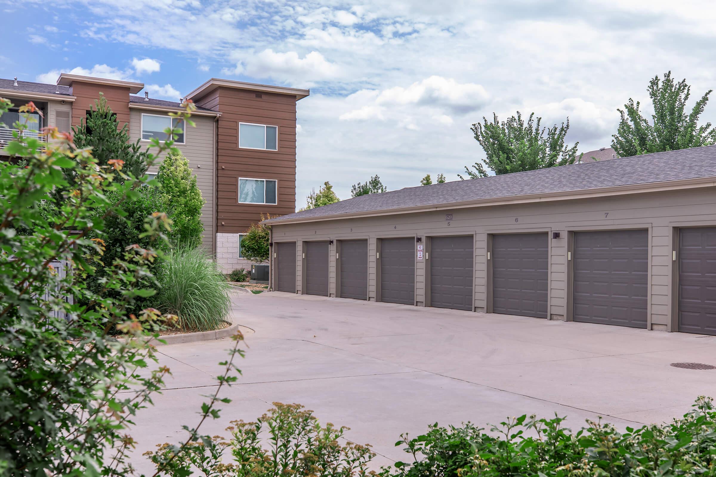 A view of a residential building with an attached garage structure. The garages feature multiple doors and are situated in a landscaped area with green shrubs and grass. The sky is partly cloudy, creating a serene ambiance.