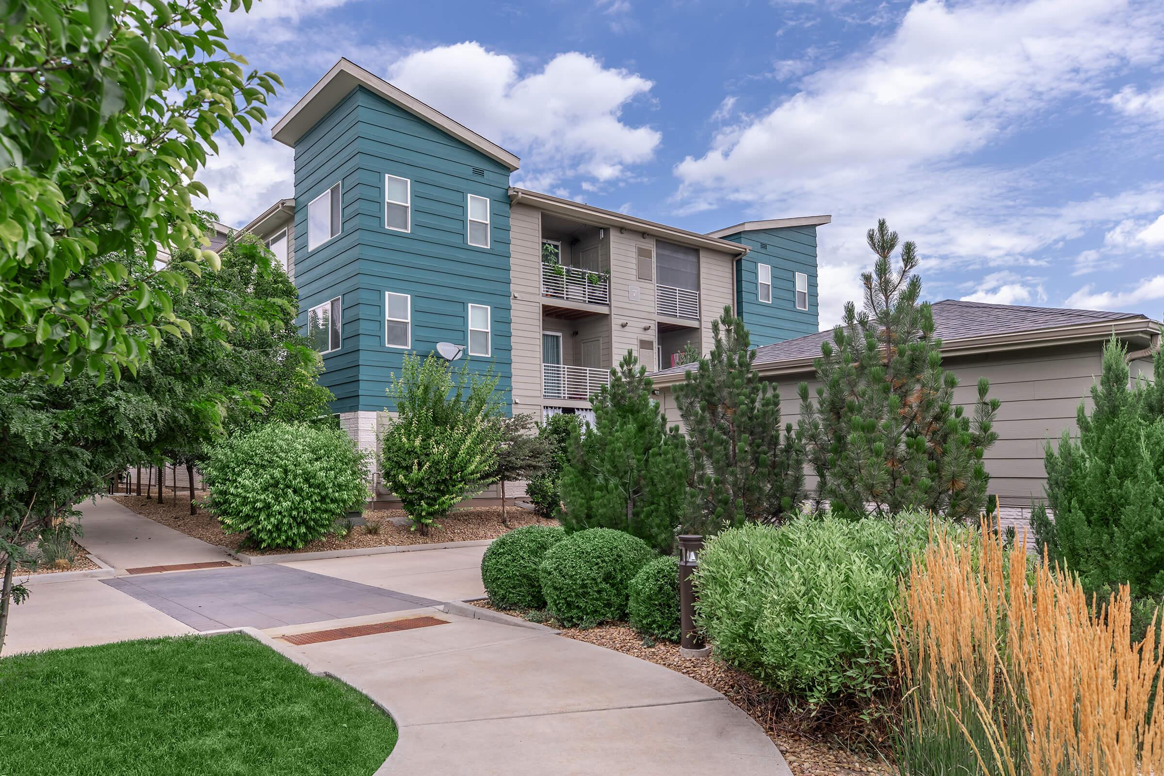 A modern multi-story residential building with a teal exterior, surrounded by lush greenery and low landscaping. A pathway leads towards the entrance, flanked by well-maintained shrubs and ornamental grasses under a partly cloudy sky.