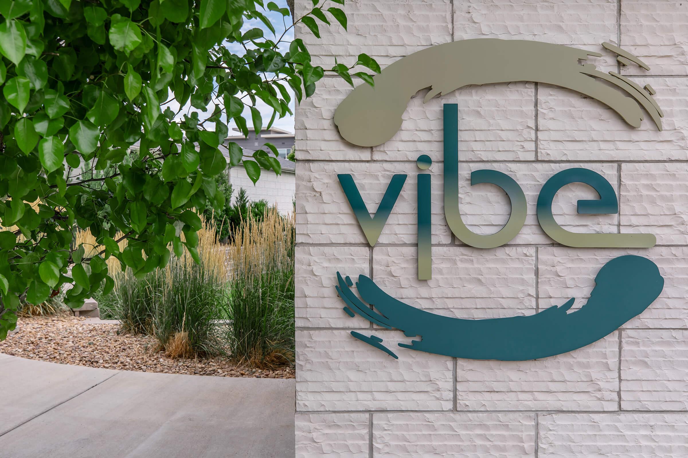 A modern sign featuring the word "vibe" in stylized letters on a textured wall. The sign is accompanied by abstract swirl designs in green and blue. In the background, there are green plants and landscaping elements, creating a welcoming atmosphere.