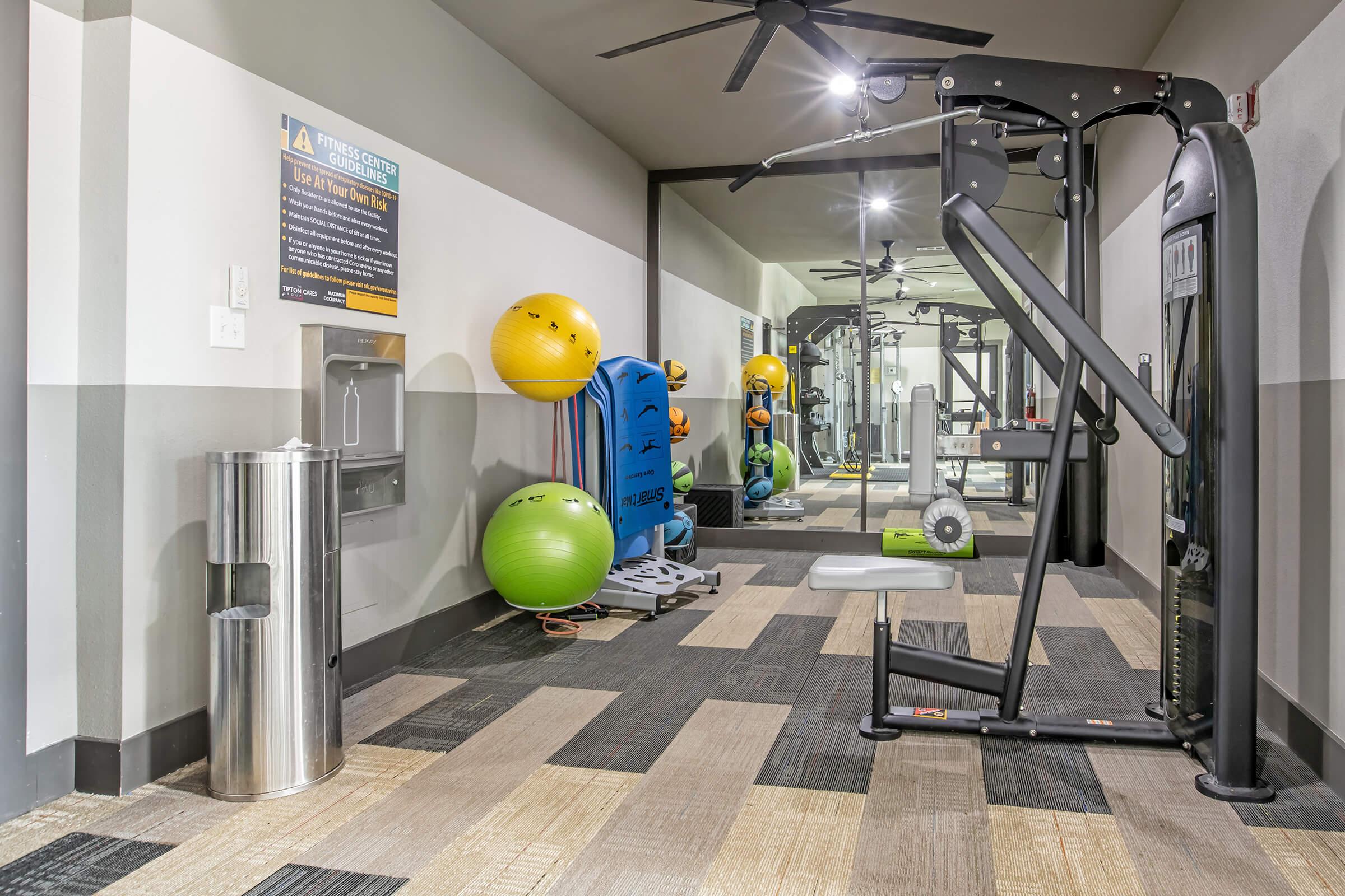 A gym area featuring exercise equipment, including a multi-use machine and workout benches. Colorful stability balls are positioned against the wall. A water fountain and a trash can are also visible in the space, with mirrors reflecting the activity area. The flooring is a mix of tiles and carpet.