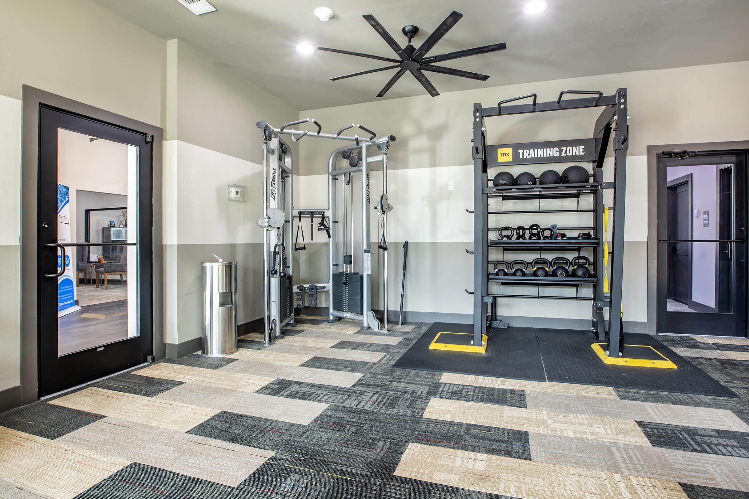 A modern gym interior featuring exercise equipment. On the right, a weight rack holds various dumbbells, while a multifunctional training machine is visible on the left. The floor is covered in patterned mats, and there are large doors and a ceiling fan, providing a spacious and well-lit workout environment.