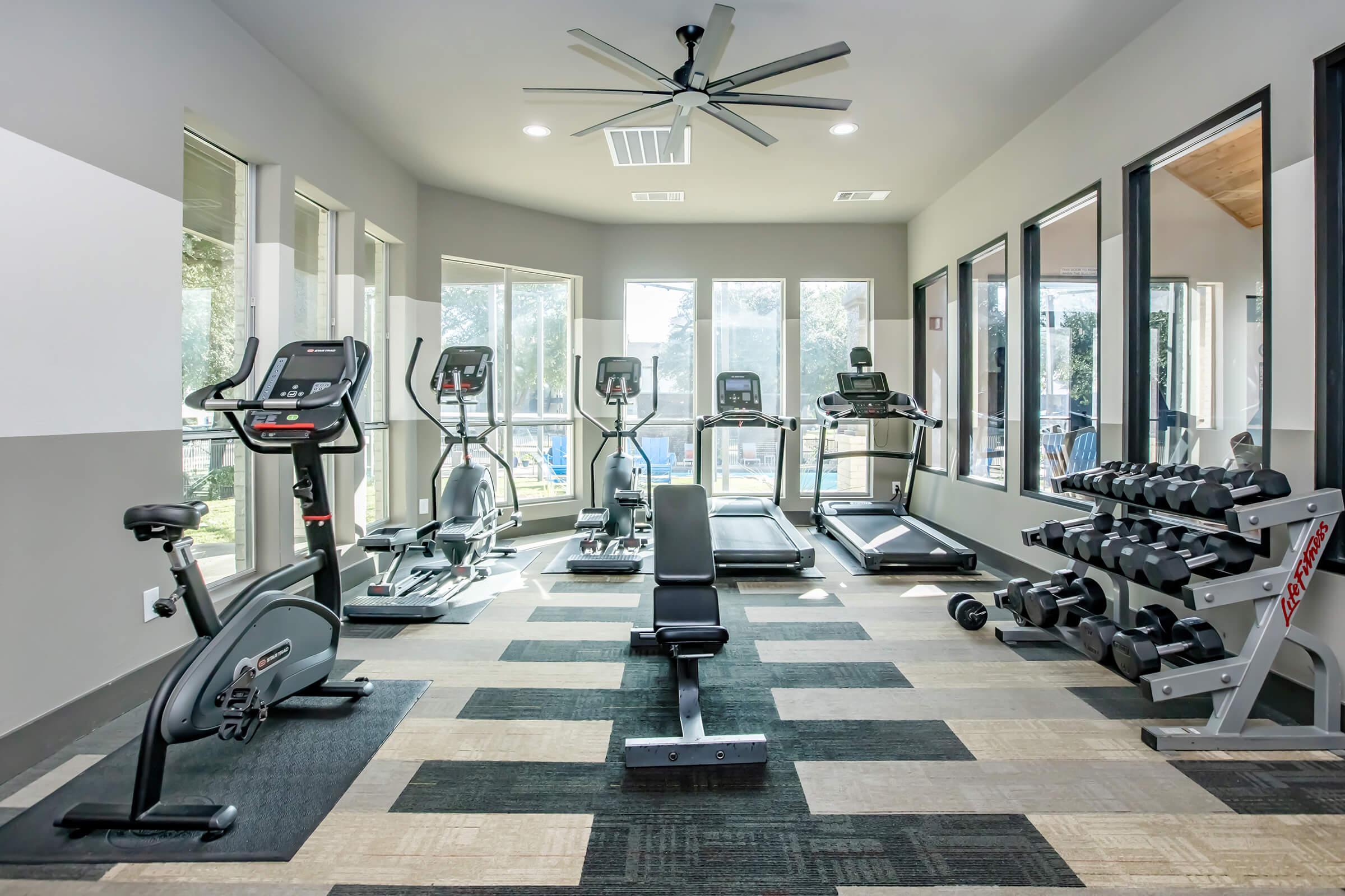 A well-lit gym interior featuring various exercise equipment including treadmills, an elliptical machine, a stationary bike, and a weight bench. Dumbbells are neatly arranged on a rack. Large windows allow natural light to fill the space, enhancing the workout environment.