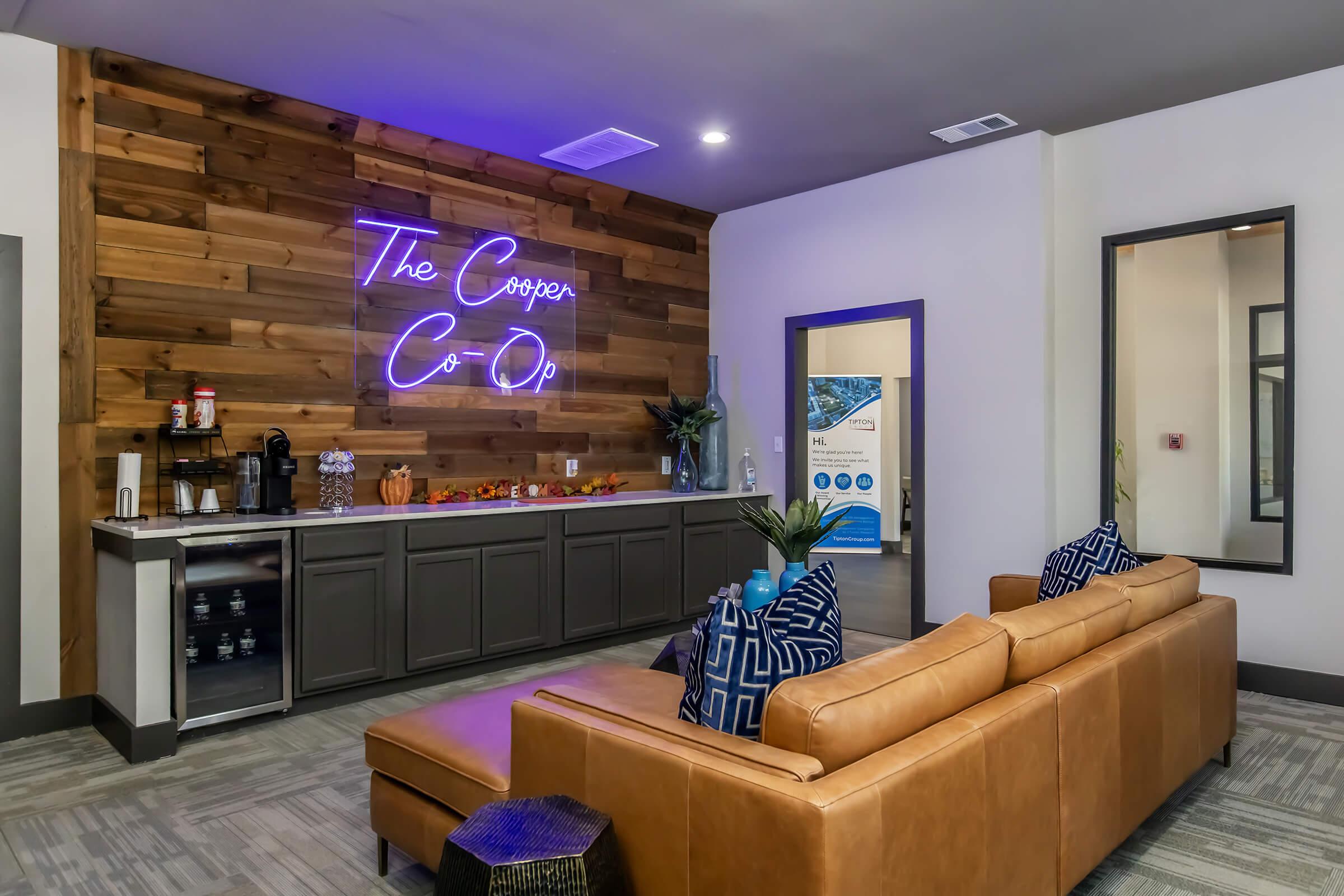 A modern lounge area featuring a wood-paneled wall with neon signage that says "The Cooper Co-Op." There’s a stylish leather sofa with decorative pillows, a side table with refreshments, and a welcoming atmosphere enhanced by indoor plants and soft lighting.