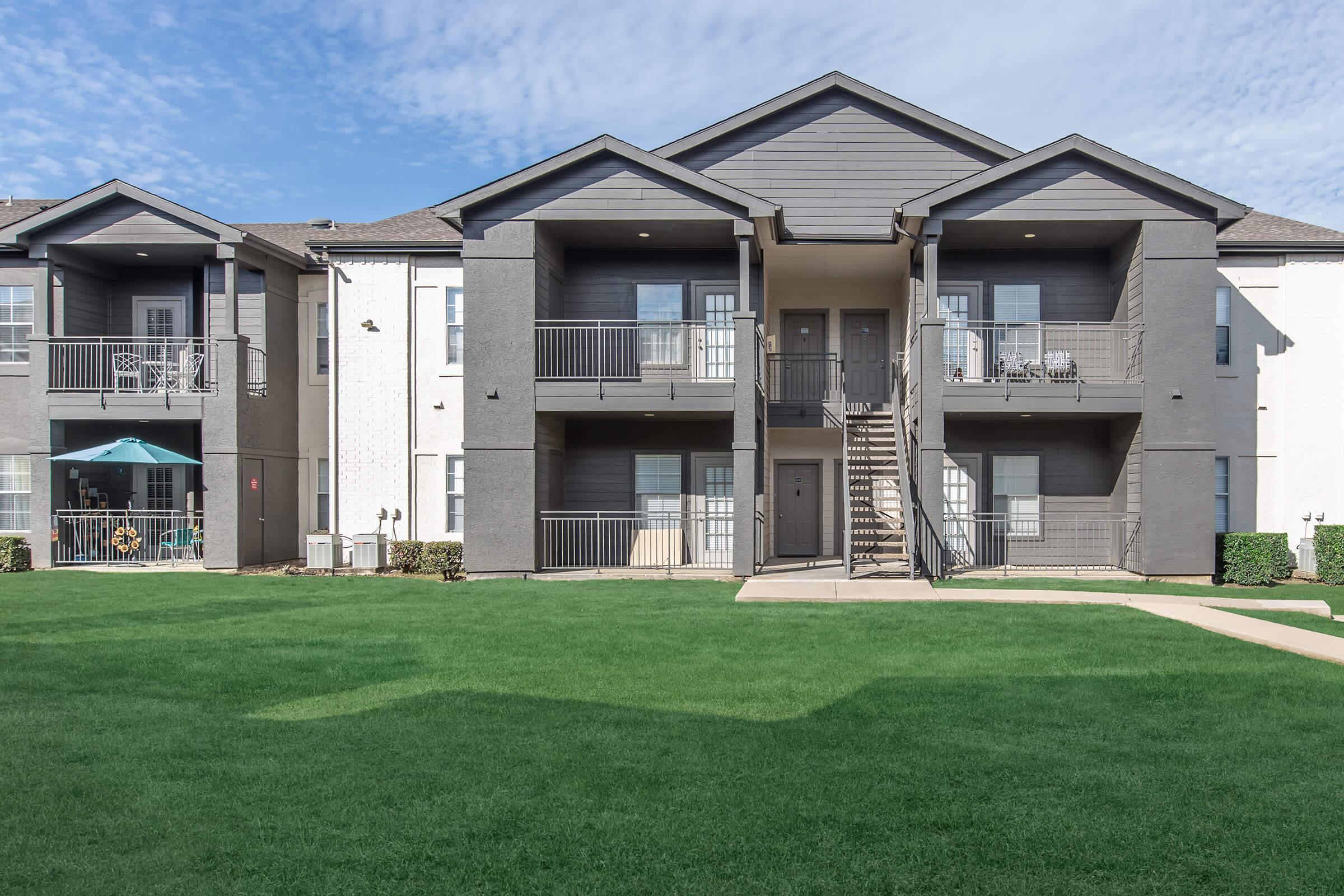 A modern two-story apartment building with a light gray and dark gray exterior. The building features balconies with black railings and a staircase leading to the second floor. Lush green grass covers the yard, and the sky is partly cloudy.