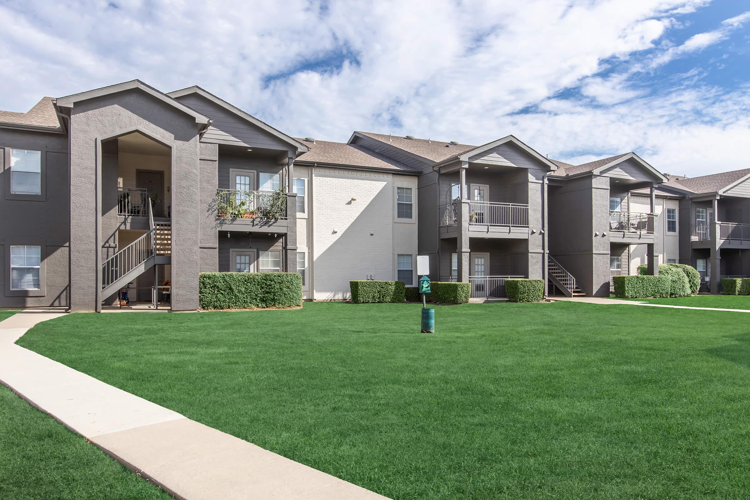 A well-maintained apartment complex featuring two-story buildings with balconies, surrounded by green lawns. Clear blue skies with some clouds create a pleasant atmosphere. A concrete walkway leads through the lawn, enhancing accessibility around the property.