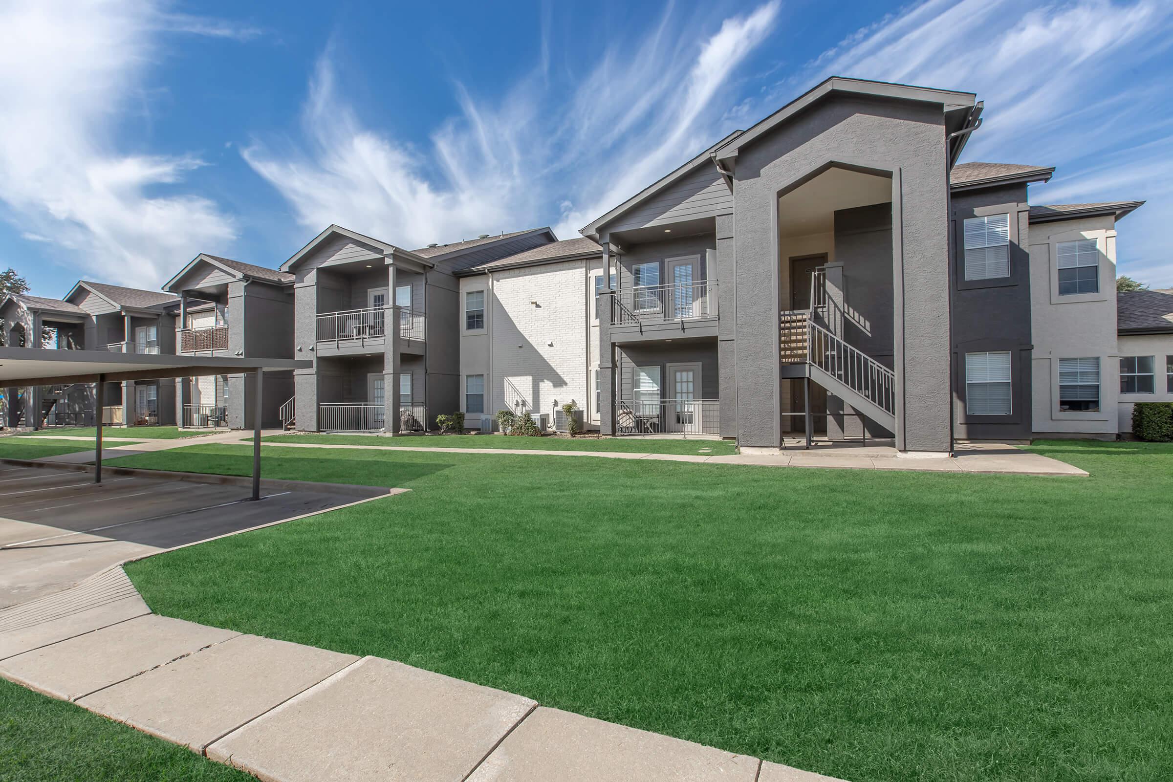 A view of a residential apartment complex featuring two-story buildings with balconies, situated on a well-maintained green lawn. The sky is bright with fluffy clouds, and a covered parking area is visible in the foreground. The modern architecture emphasizes a cozy community atmosphere.