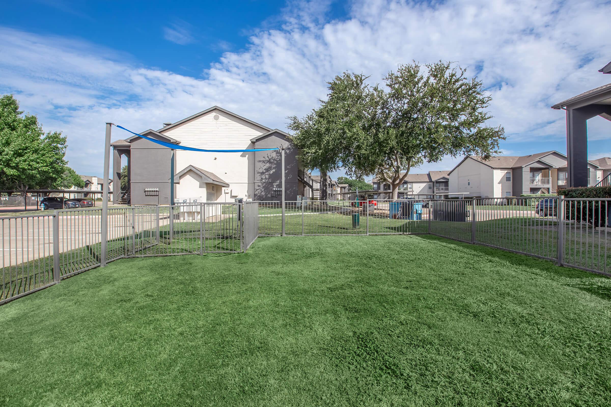 A fenced grassy area with a few trees and a light-colored building in the background. The sky is partly cloudy, and there are other buildings visible in the distance. The space appears well-maintained and is likely designed for recreational use.
