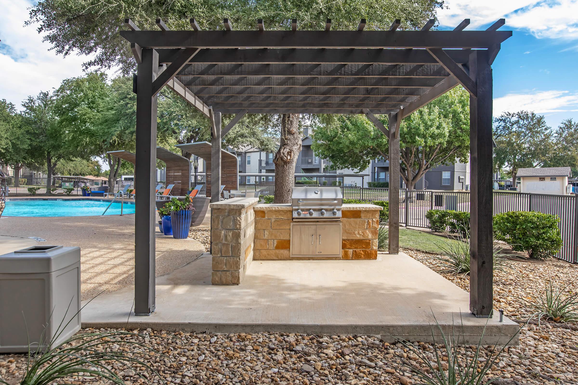 Outdoor grilling area with a stone barbecue under a wooden pergola. Nearby is a swimming pool surrounded by lush greenery and lounge chairs. The setting is bright and inviting, perfect for gatherings and outdoor cooking.