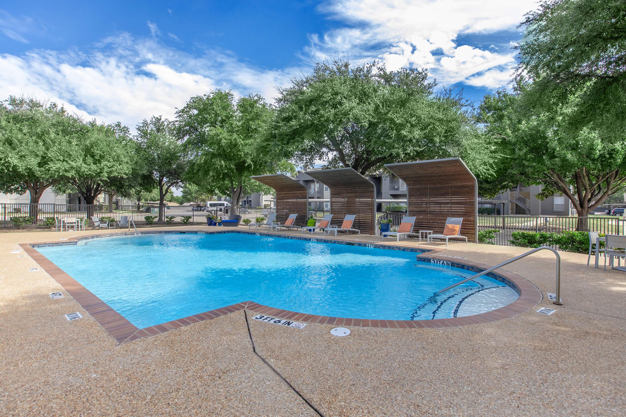 A refreshing swimming pool surrounded by a patio with lounge chairs and shaded cabanas. Lush green trees line the area under a bright blue sky with scattered clouds, creating a relaxing outdoor atmosphere.