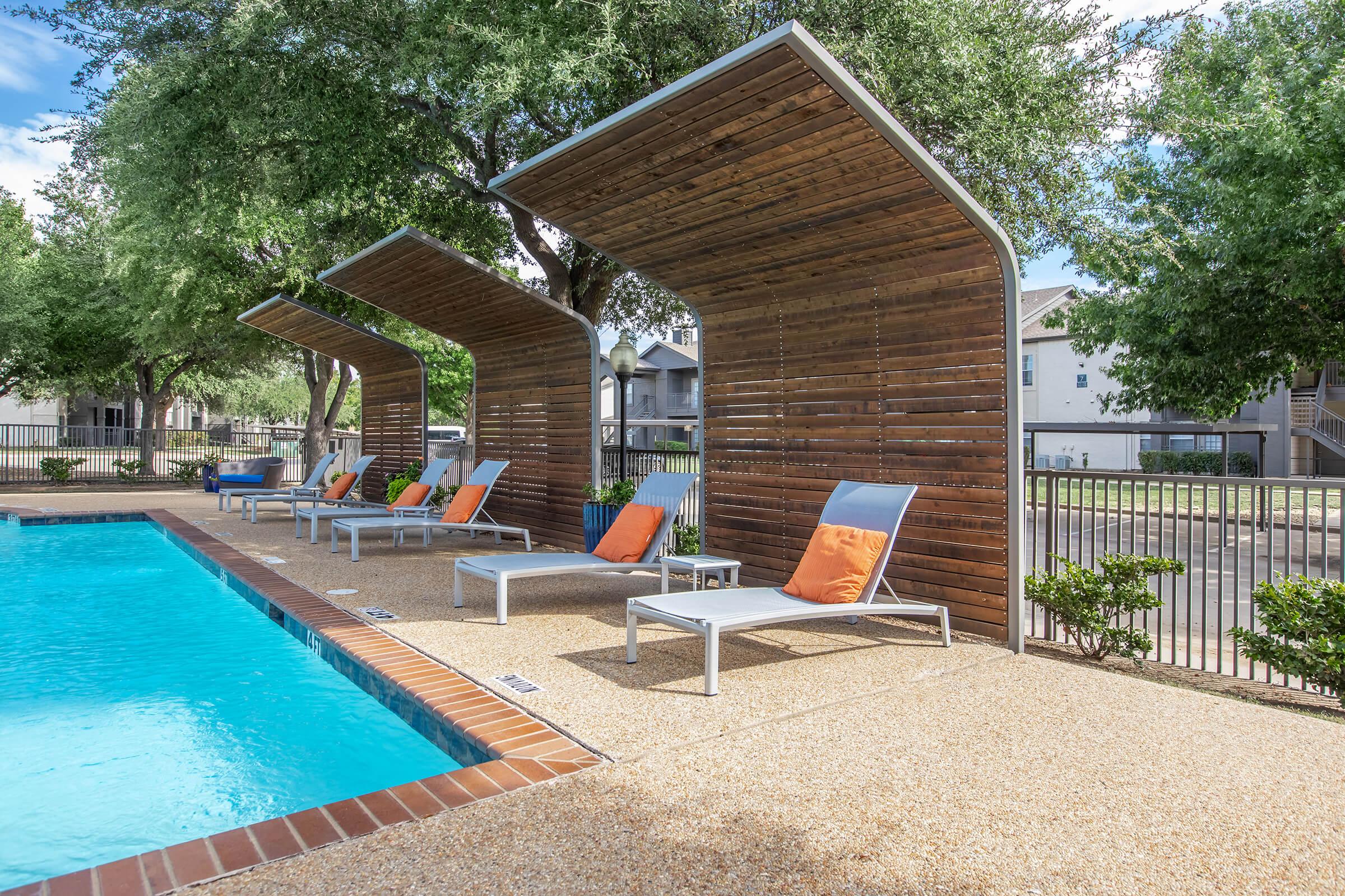 A serene poolside area featuring three lounge chairs with orange cushions under wooden canopies. Lush green trees line the background, and a well-maintained pool reflects the clear sky in front. The setting is inviting and perfect for relaxation.
