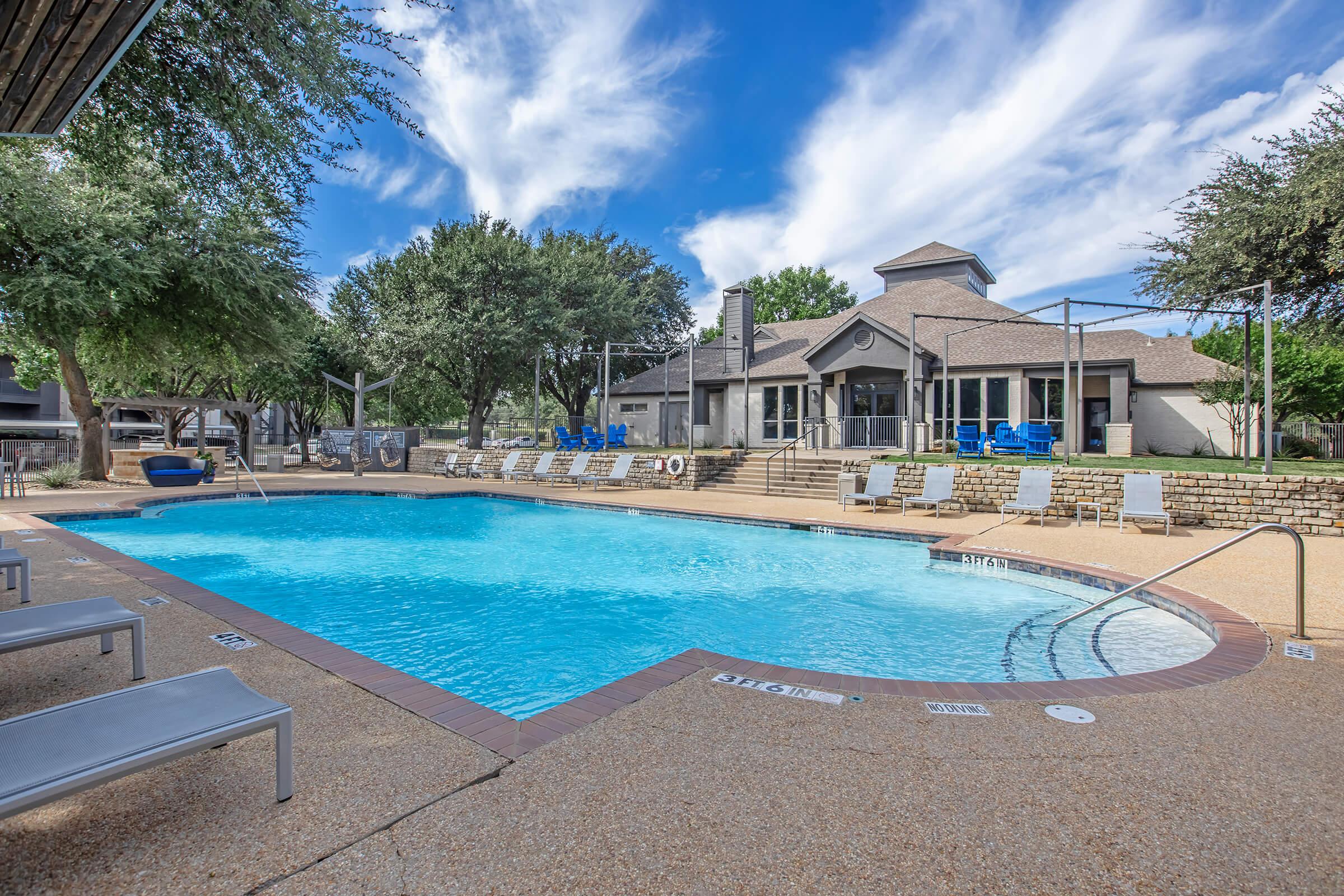 A swimming pool area surrounded by lounge chairs, with a spacious deck and a clubhouse in the background. Lush green trees and a blue sky with clouds provide a relaxing atmosphere. The pool has a gentle curve and a shallow entry, inviting visitors to enjoy a refreshing swim.