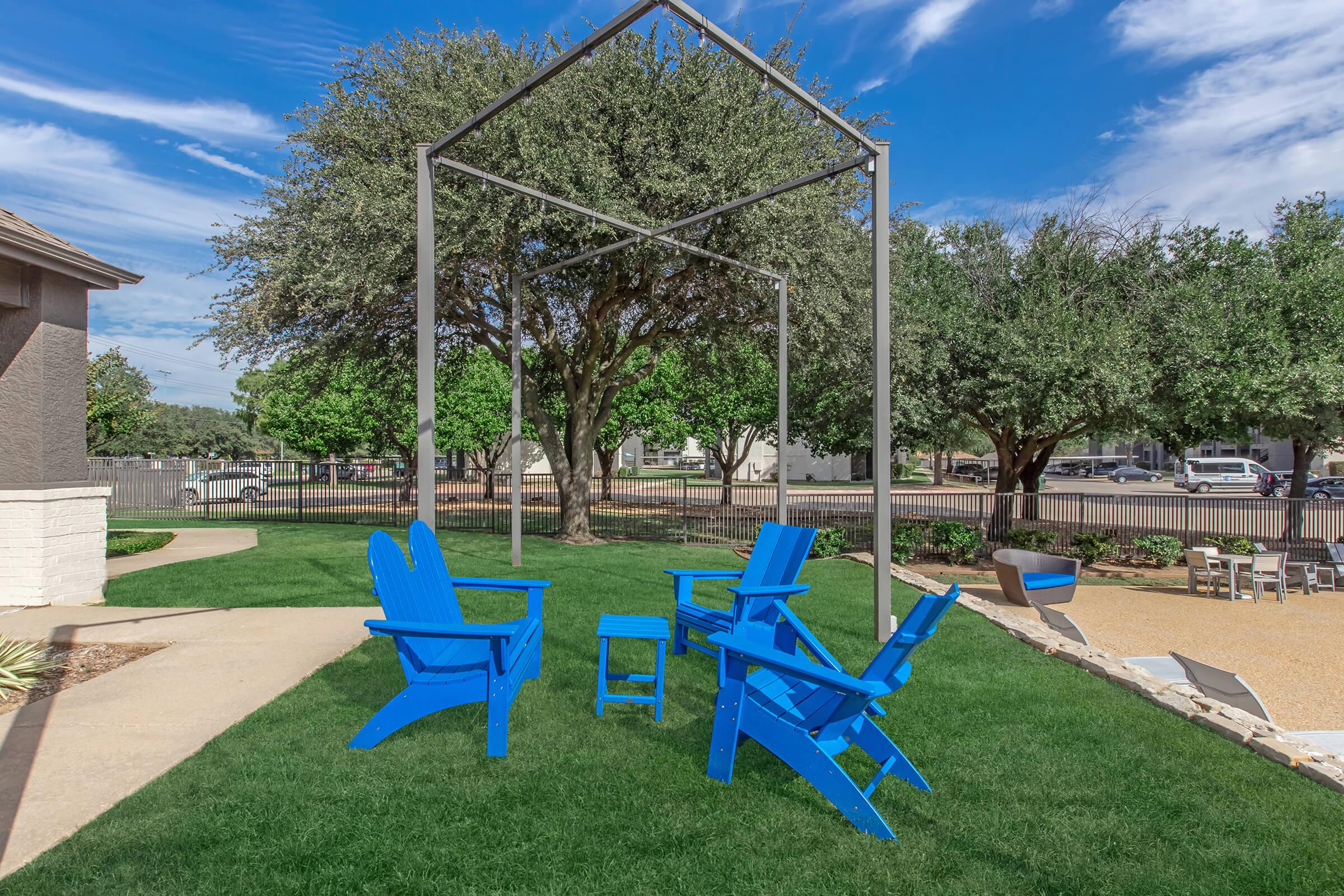 A serene outdoor space featuring vibrant blue Adirondack chairs surrounding a small table under a modern metal frame structure. Lush green grass and well-maintained trees provide a calming ambiance, with a pathway leading to the seating area and a gravel section nearby.