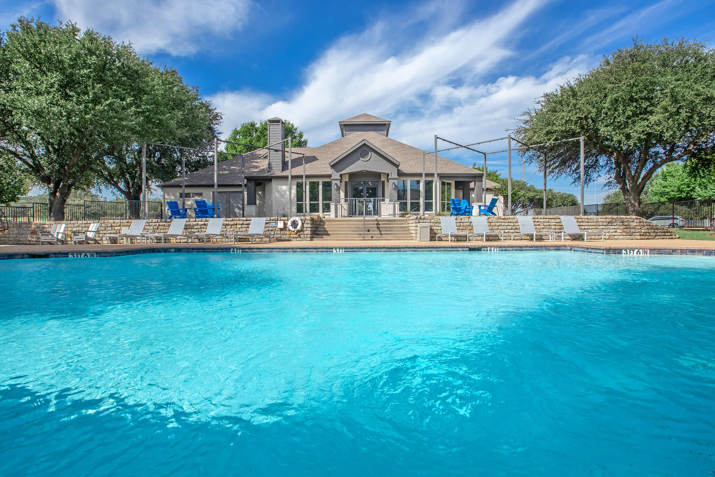 A scenic view of a swimming pool with clear blue water, surrounded by lounge chairs. In the background, a modern building with large windows and a shaded porch is visible, along with trees and a bright blue sky scattered with clouds. The area appears inviting and well-maintained.