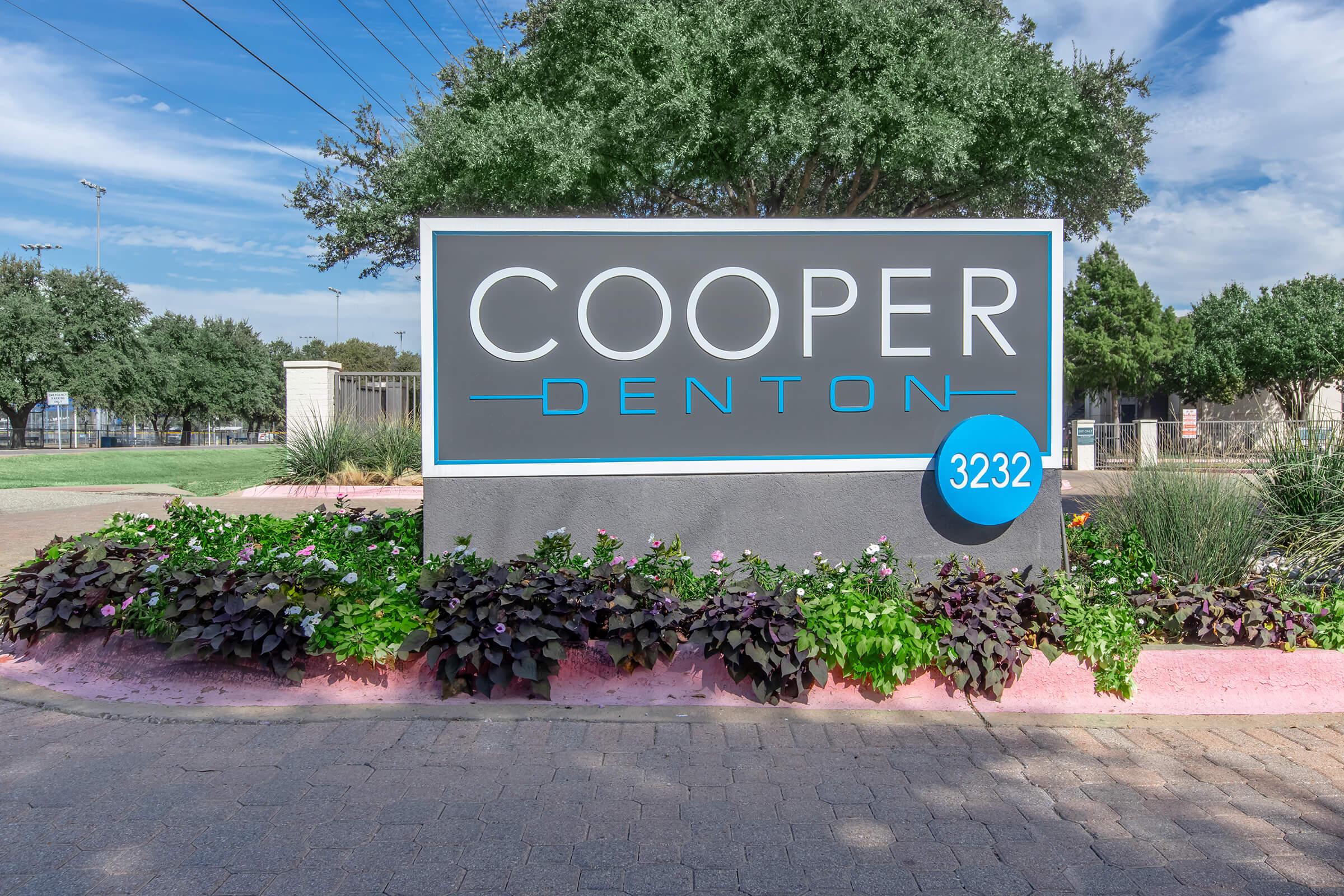 Sign for "Cooper Denton" located at 3232, surrounded by green landscaping and colorful flowers. The sign features a modern design with a dark background and light blue lettering, set against a clear blue sky.
