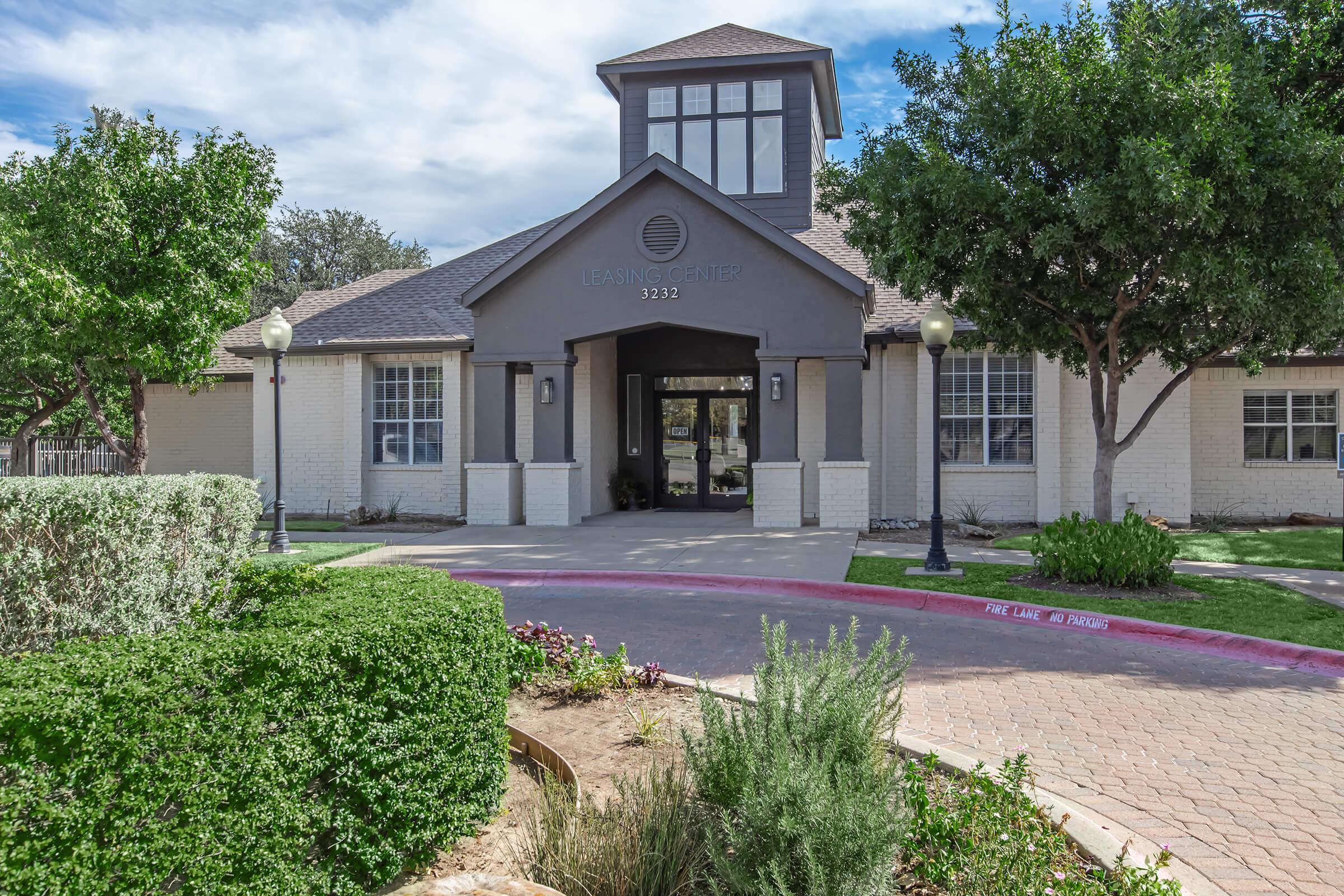 A modern building with a peaked roof and a central entrance, surrounded by neatly trimmed landscaping. The structure features light and dark grey walls, large windows, and decorative lampposts. Trees and shrubs add greenery to the entrance area, and a paved path leads up to the doorway.