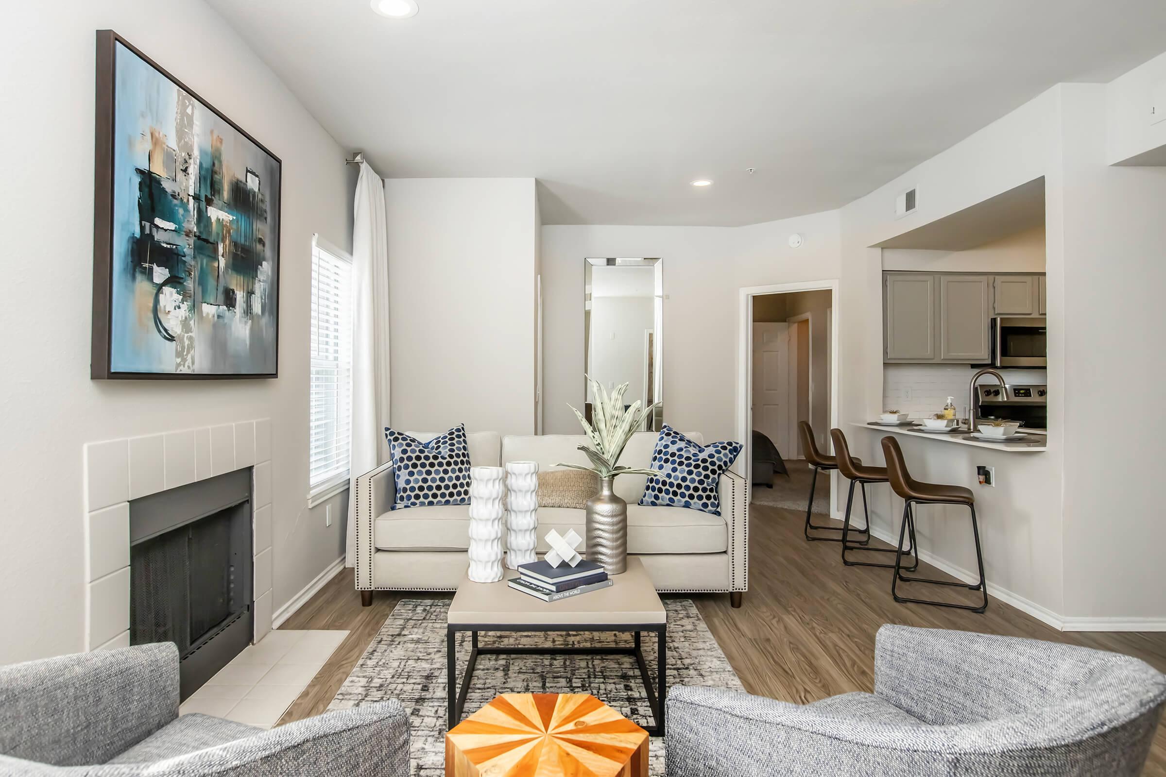 Living room featuring a cozy sofa with decorative pillows, two stylish chairs, a coffee table, and a modern art piece on the wall. The room has a fireplace, a large window with sheer curtains, and an open layout leading to a kitchen area with bar stools. Neutral color palette and contemporary decor create a welcoming atmosphere.