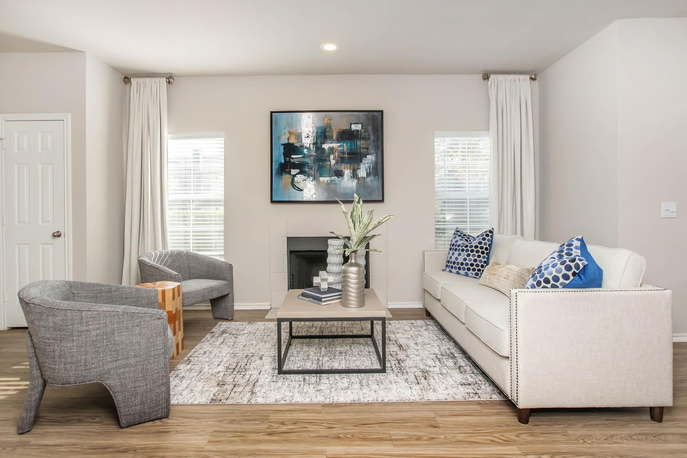 A modern living room featuring a white sofa with blue and white patterned cushions, two gray accent chairs, a wooden side table, and a coffee table with vases. Light-colored walls, large windows with curtains, and an abstract painting on the wall create a bright, inviting atmosphere.