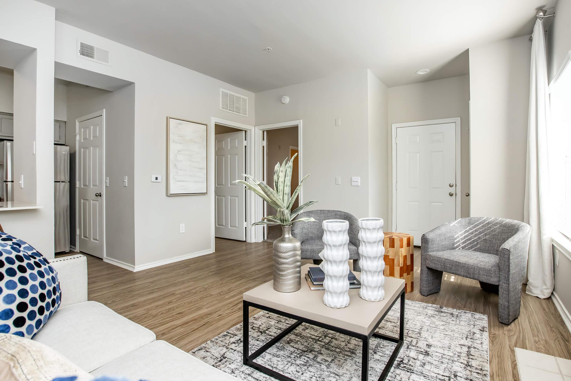 A modern living room featuring a light gray couch with blue patterned pillows, a coffee table with decorative vases, and a small plant. The space has light-colored walls, wooden flooring, and two armchairs. There are doors leading to other rooms, adding to the inviting atmosphere.