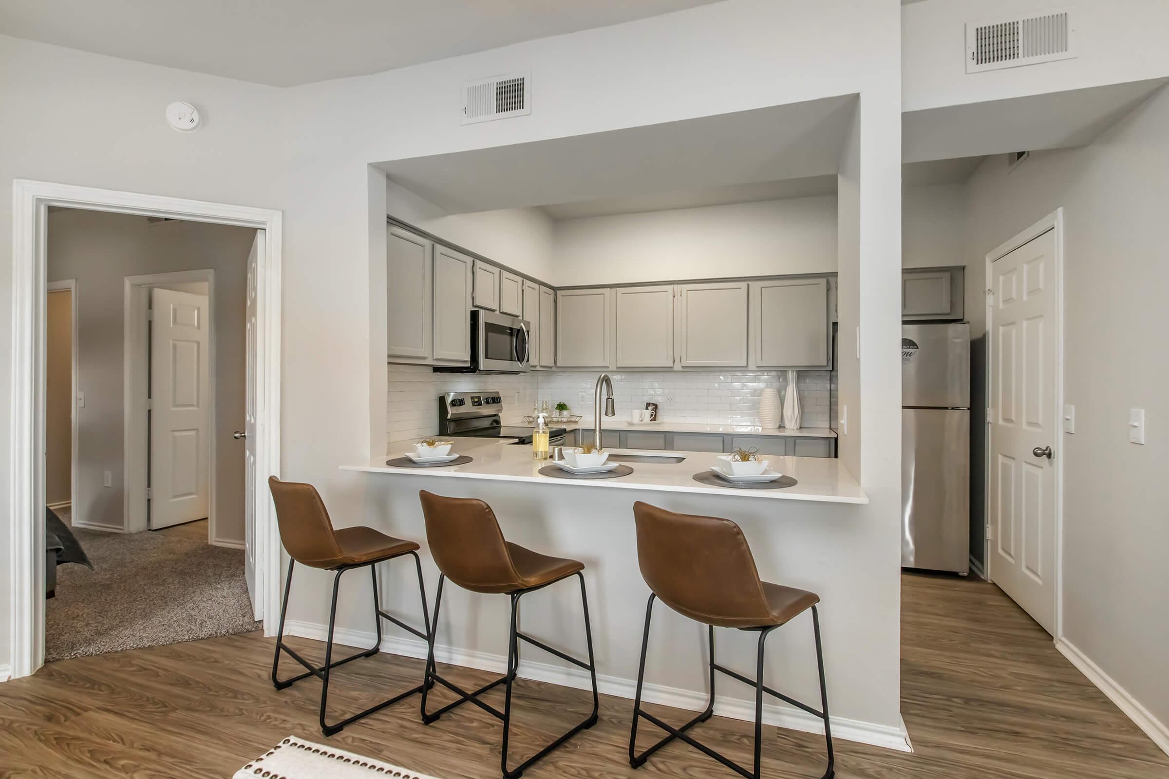 Modern kitchen area with gray cabinets and stainless steel appliances. Three stylish brown bar stools are positioned at a countertop. The space features light-colored walls and wooden flooring, adding a warm ambiance. In the background, a door leads to another room, enhancing the open-concept design.