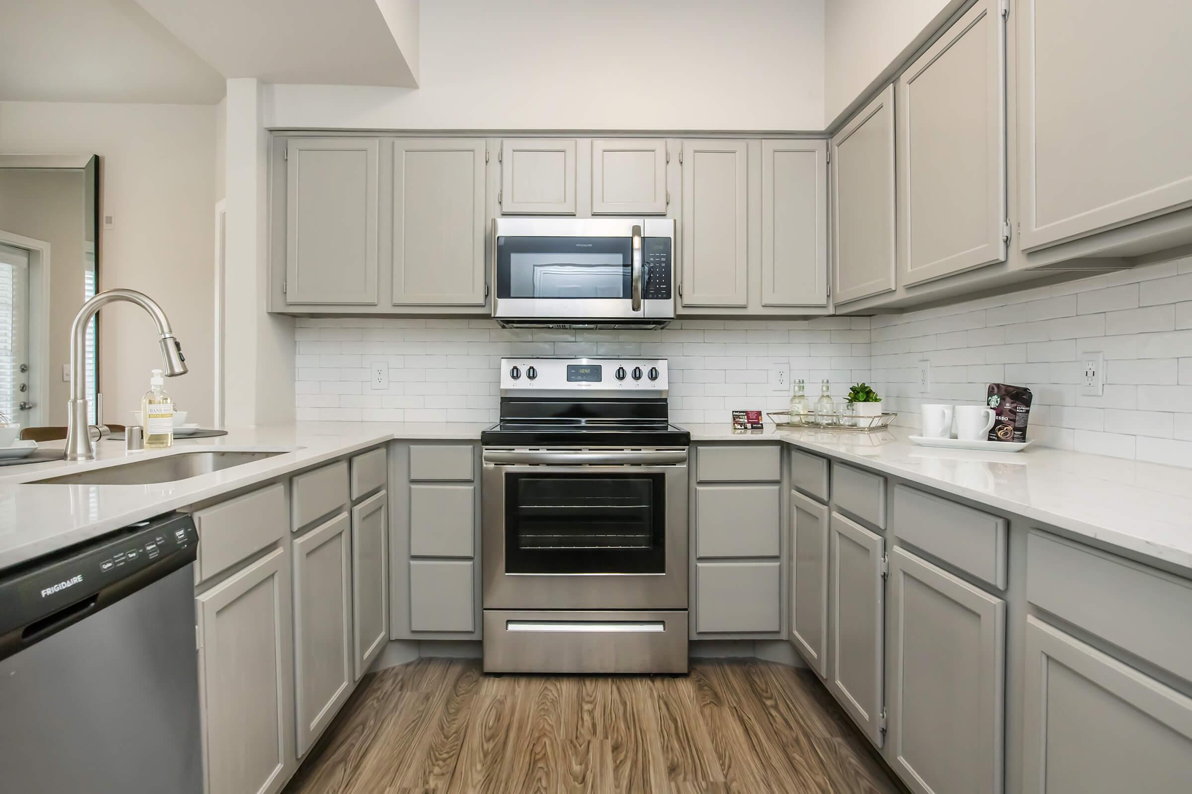 Modern kitchen featuring gray cabinets, sleek stainless steel appliances, including a built-in microwave and stove. The countertops are light-colored, complemented by a white subway tile backsplash. The flooring is a warm wood tone, creating a cozy atmosphere. 