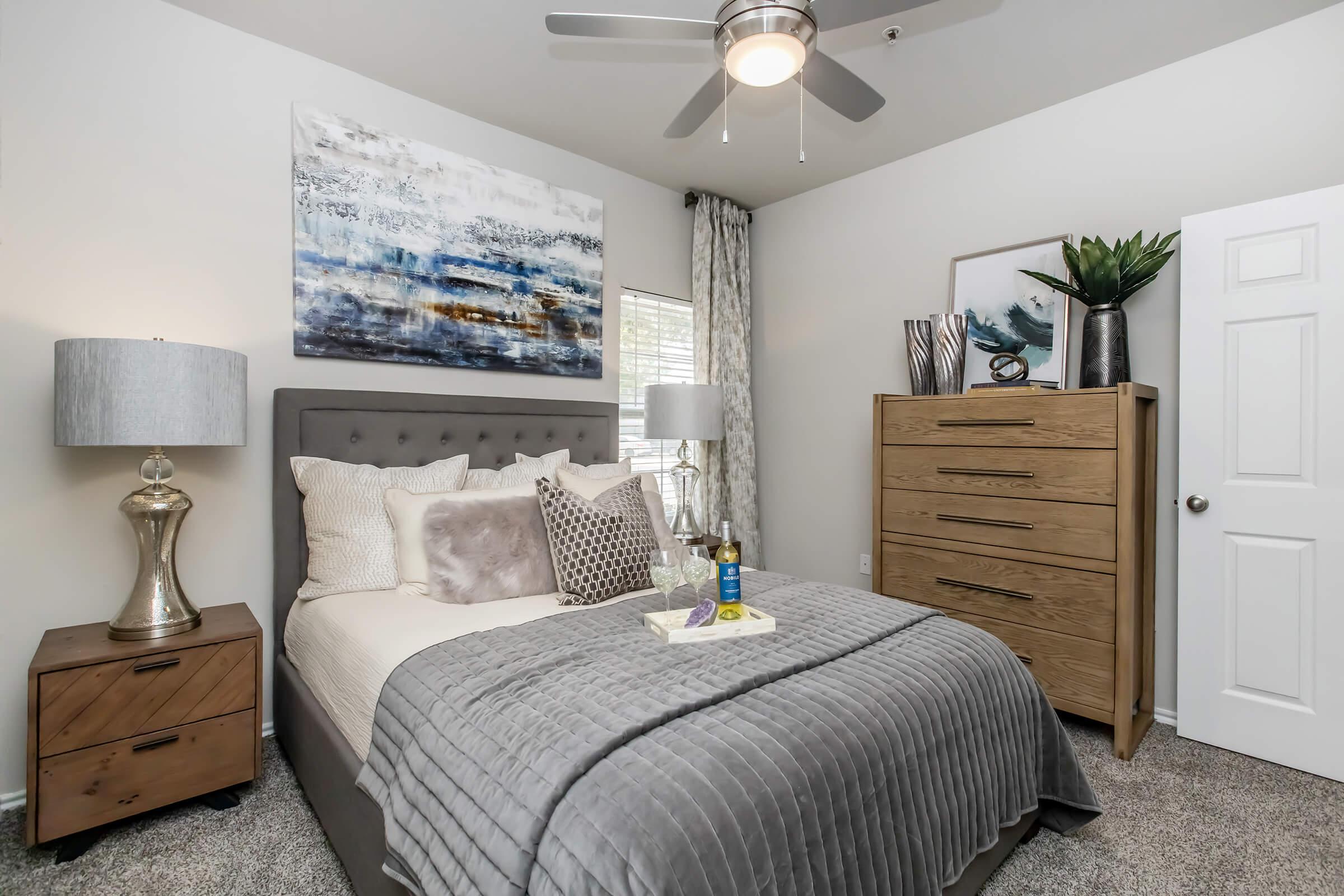 A well-decorated bedroom featuring a plush gray bedspread, decorative pillows, bedside tables with lamps, a tall wooden dresser, and artwork on the walls. A ceiling fan adds to the ambiance, and natural light filters through a window with patterned curtains. The cozy decor creates a welcoming atmosphere.