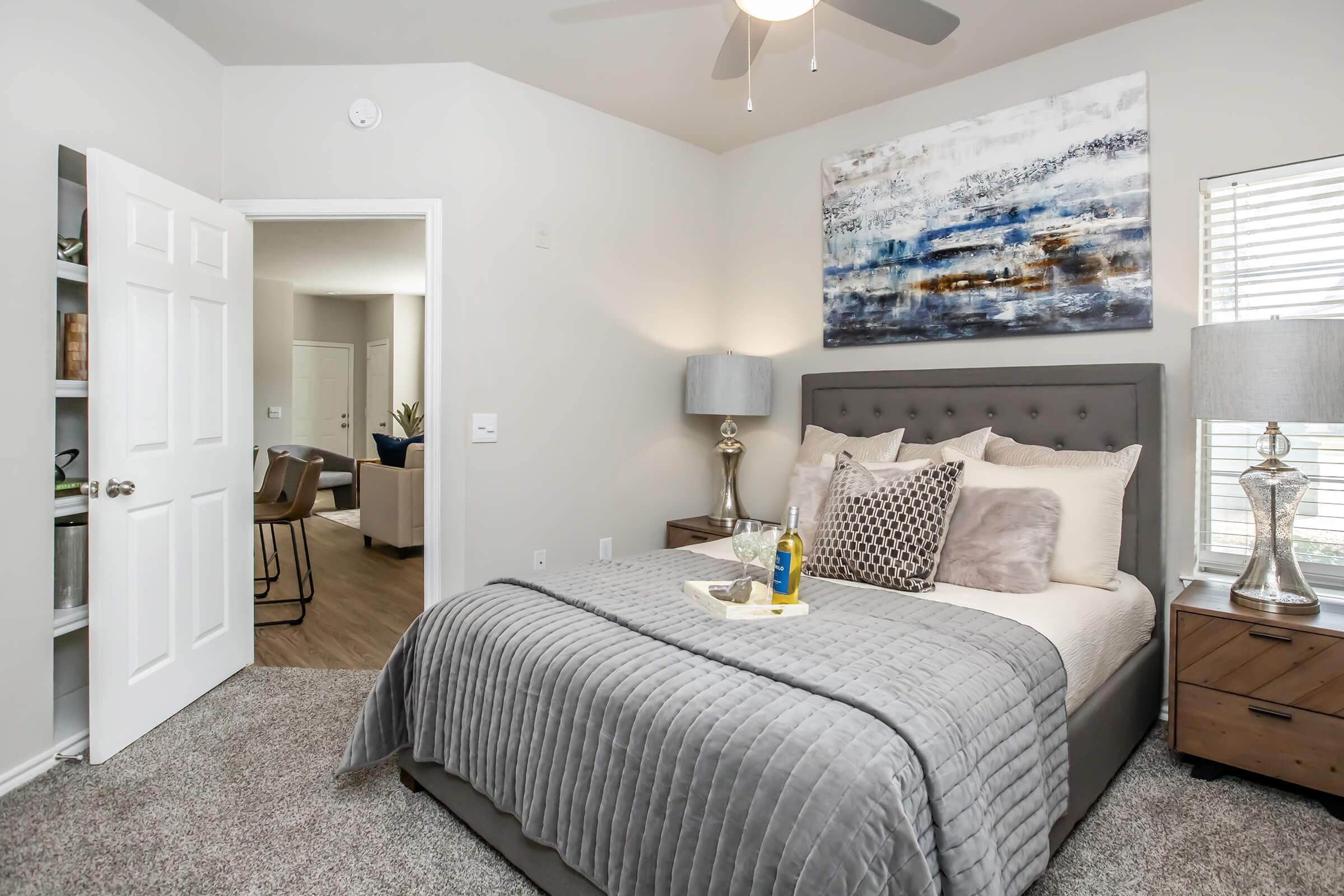 A cozy bedroom featuring a gray upholstered bed with decorative pillows and a textured throw blanket. Two bedside lamps illuminate the space, and a colorful abstract painting hangs above the headboard. A doorway in the background leads to a living area with modern furniture.