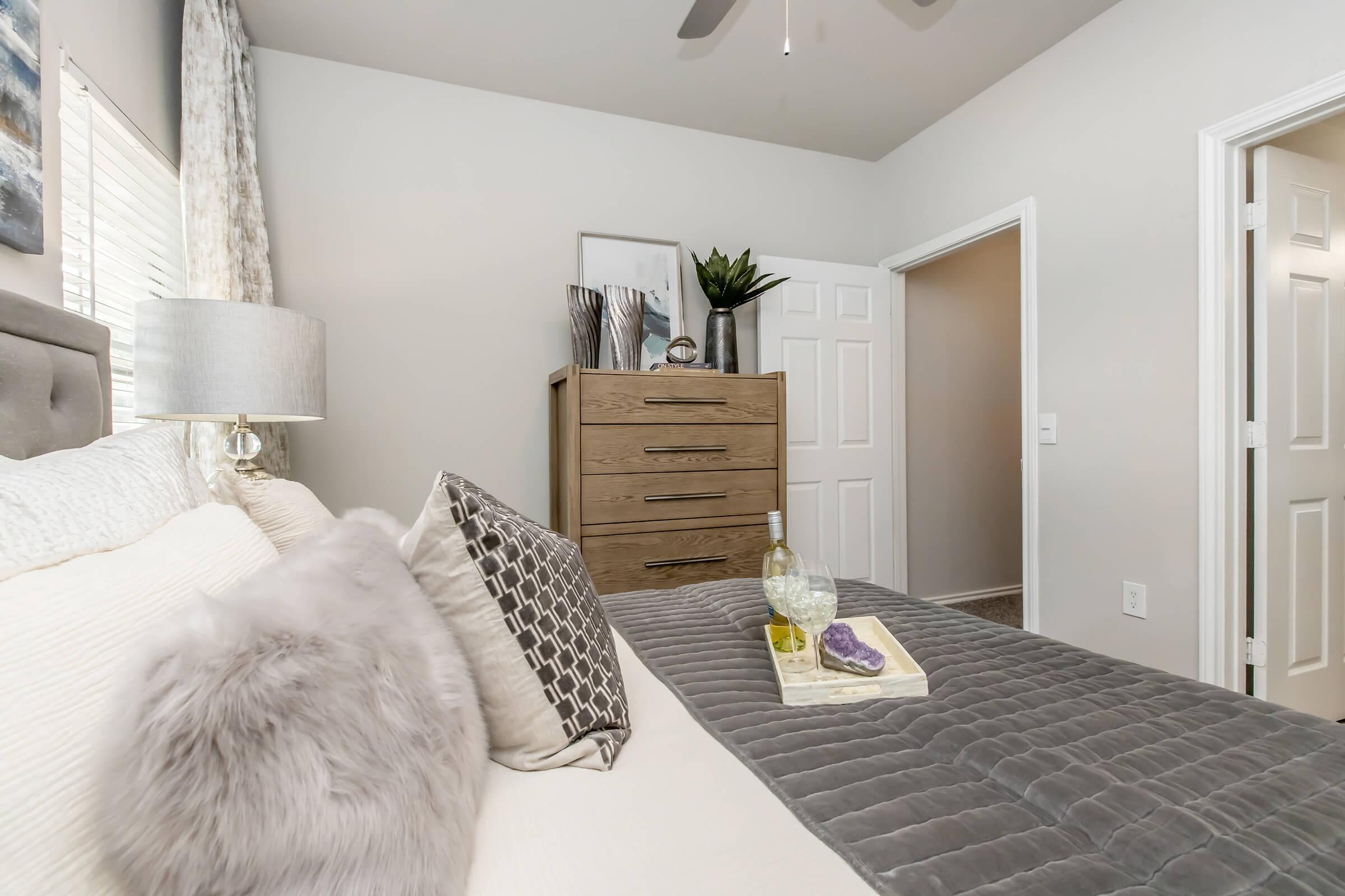 A cozy bedroom featuring a neatly made bed with decorative pillows and a gray blanket. A wooden dresser stands against the wall, adorned with a plant and decorative items. Light streams in through a window, and there is an open door leading to another room, creating a welcoming atmosphere.
