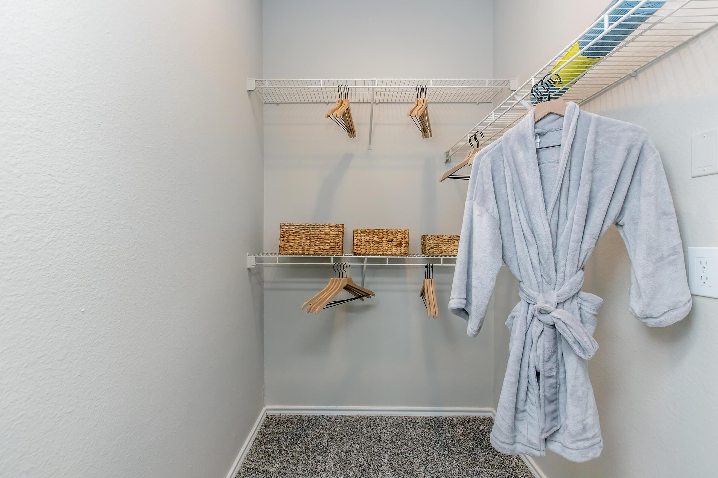 A neatly organized closet with gray walls. It features wire shelves holding several wooden hangers and two woven baskets. A soft, gray robe hangs on the side, adding a cozy touch to the space. The floor is covered in dark carpet, enhancing the minimalist design.